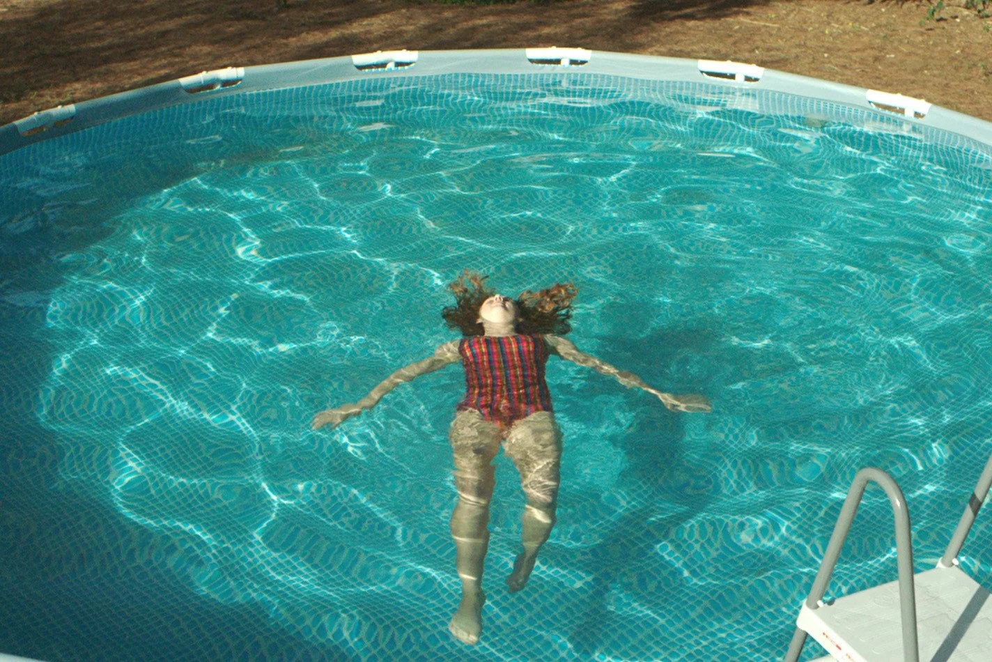 A woman is floating on her back in an above-ground swimming pool with her arms outstretched and eyes closed.