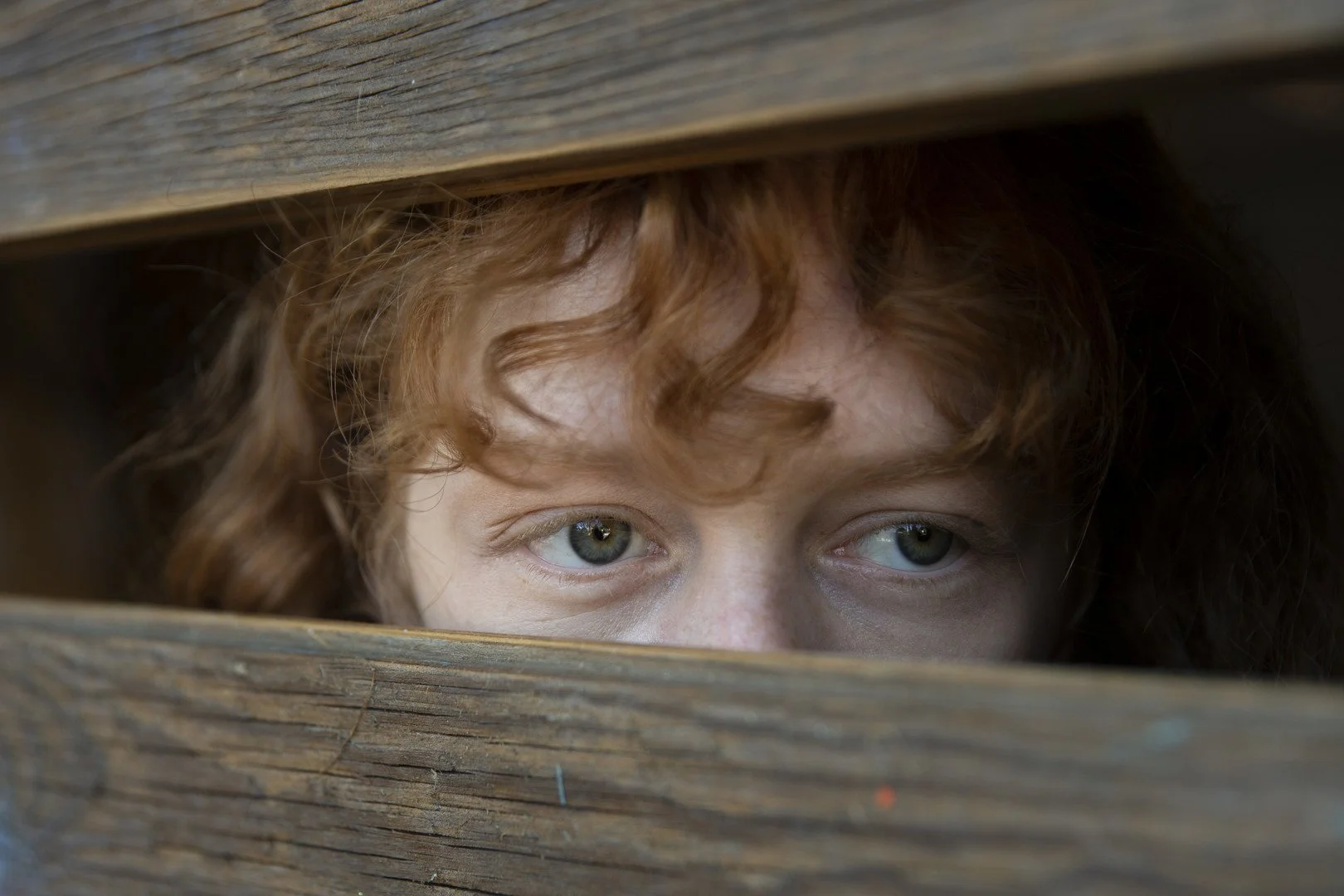 Close-up of a person's face with curly red hair peeking through a gap in wooden planks, showing their blue eyes.