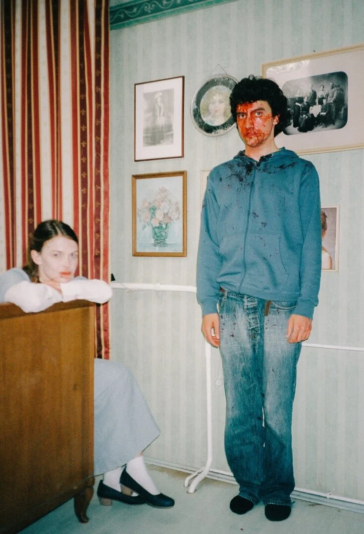 A young woman sitting at a desk with her head resting on her arms, looking at the camera, and a young man standing beside her with face covered in fake blood and wearing casual clothes, in a room with framed pictures on the wall.