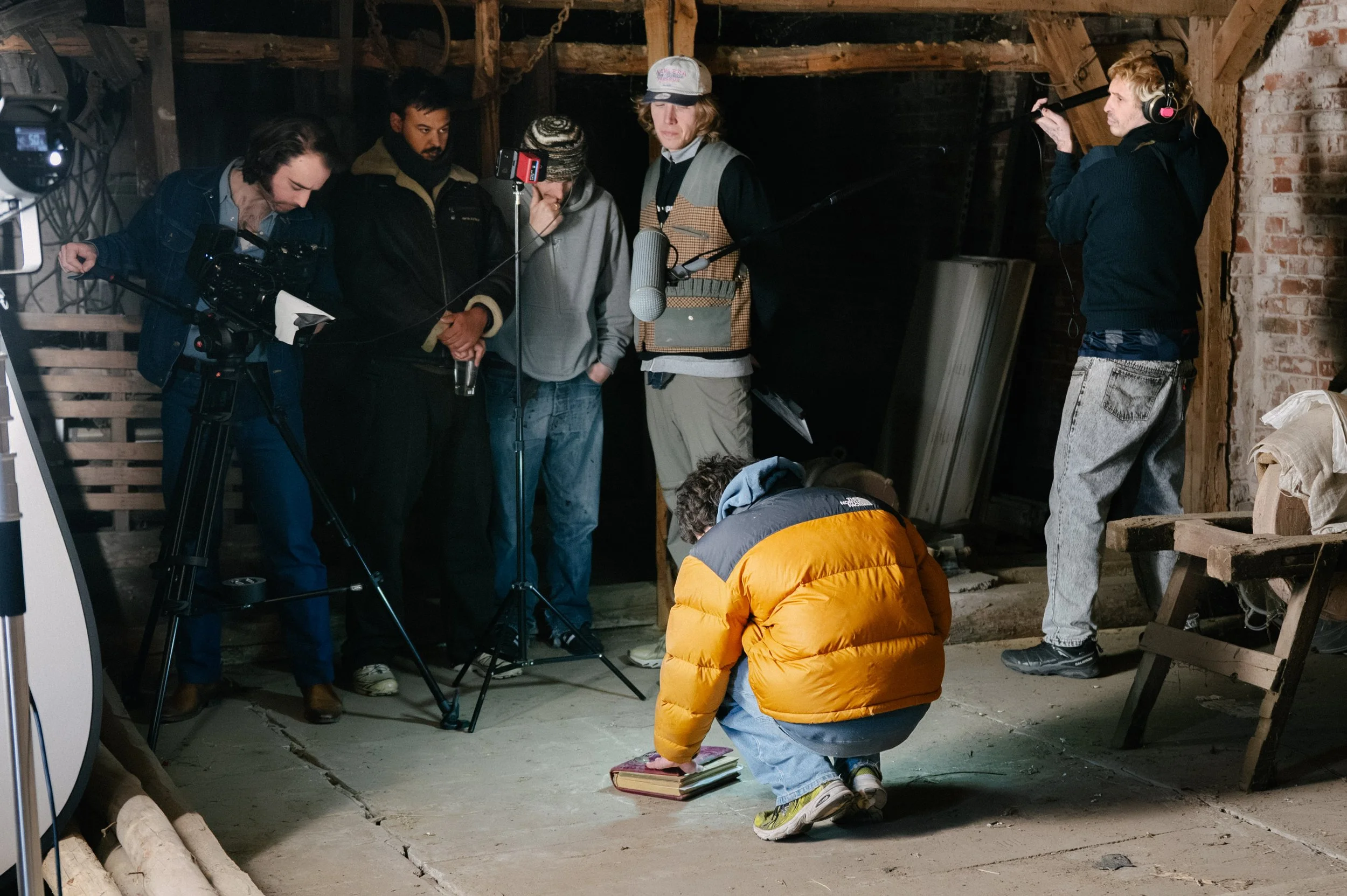 A film crew working in an attic with exposed wooden beams and brick walls. Several crew members are focused on a scene involving a child wearing a yellow puffer jacket and gray pants, who is kneeling on a stack of books. A young woman is adjusting a 