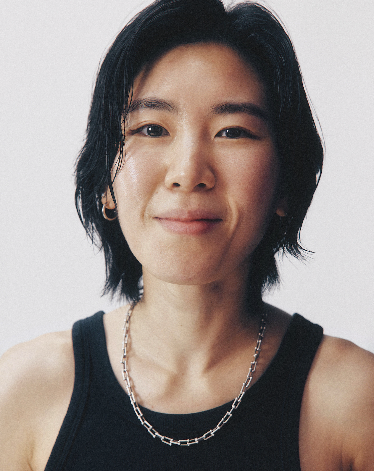 Close-up portrait of a woman with short black hair, wearing a black tank top, silver chain necklace, and small hoop earrings, smiling subtly against a plain white background.