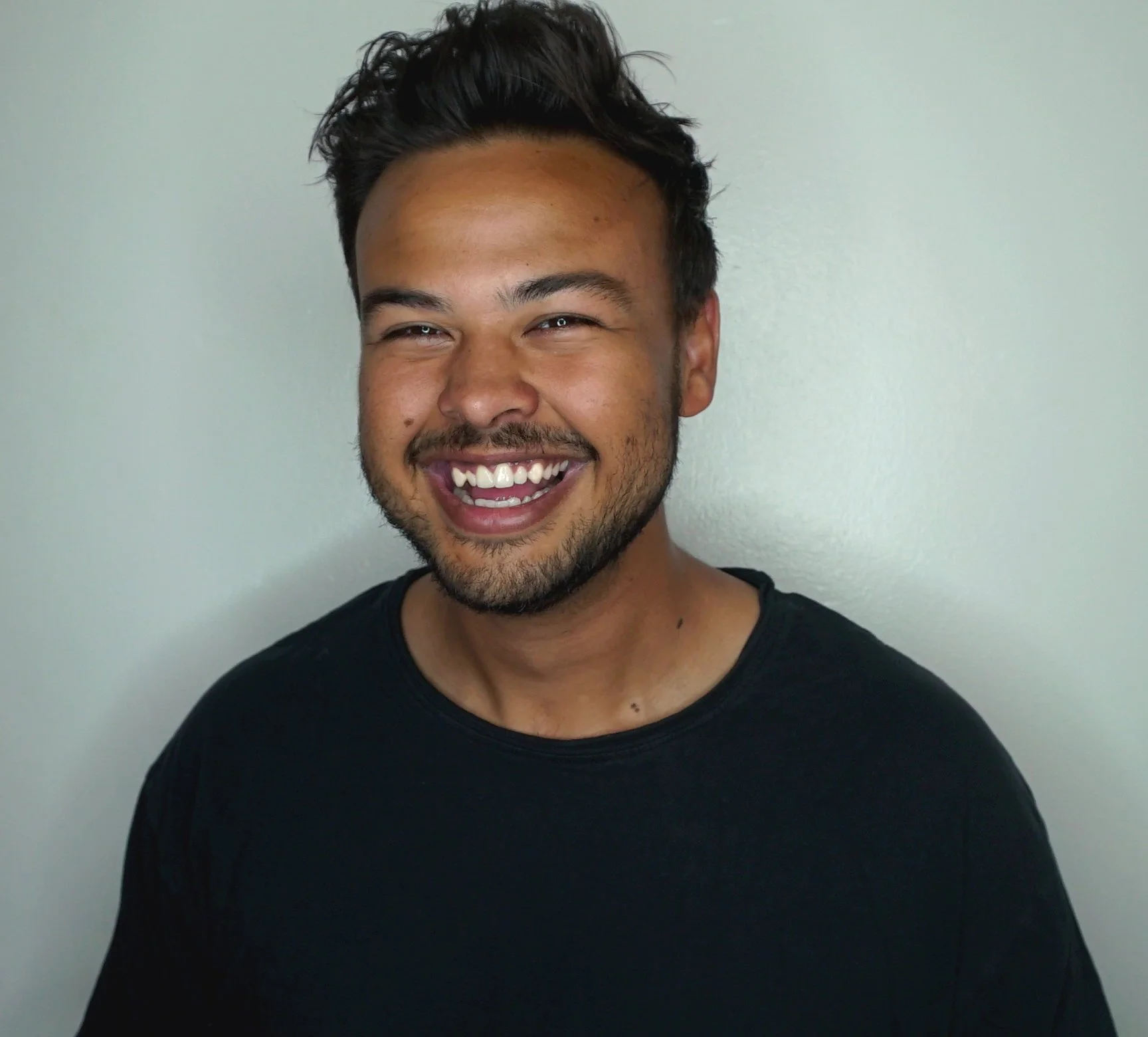 A young man with dark, tousled hair and a beard, smiling broadly, wearing a black T-shirt, against a plain, light-colored background.