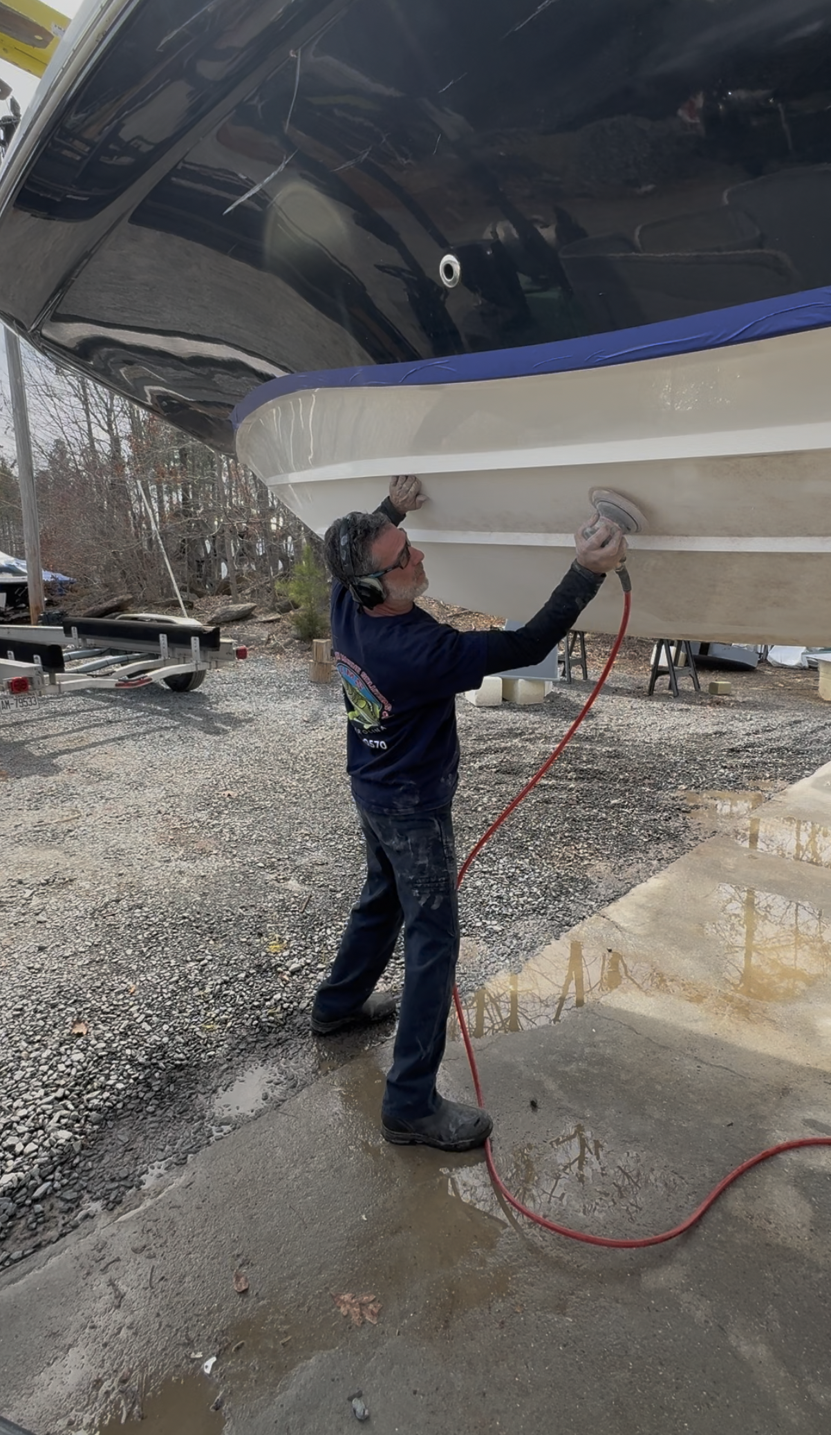 A man using a power sander on the bottom of a boat that is raised on land. The man is wearing protective eyewear and headphones, standing on a wet concrete surface.