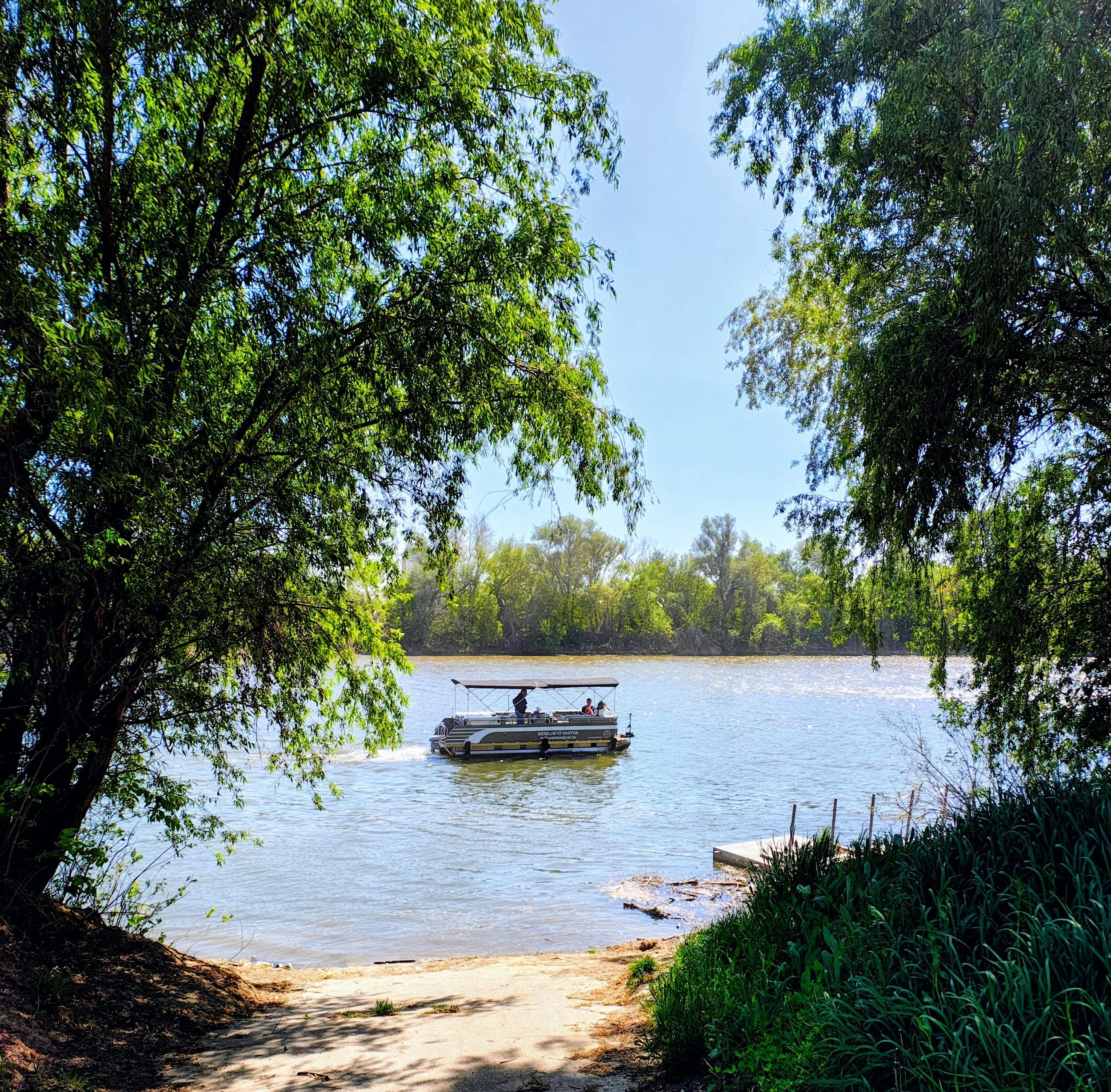A boat on a river viewed from a shaded riverbank with trees and lush greenery on both sides.