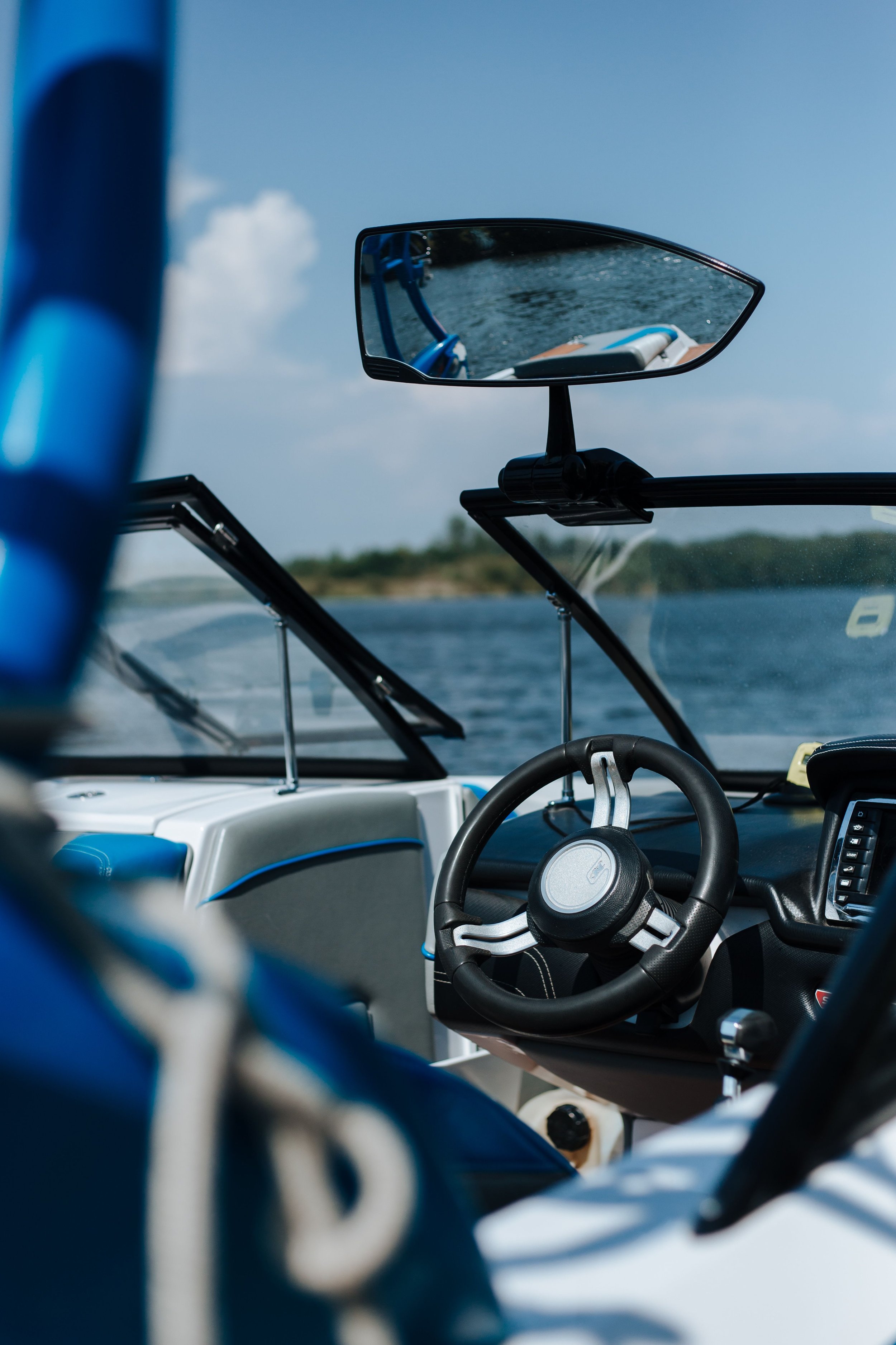 View from inside a boat showing a rearview mirror reflecting water and part of the boat, with blue sky and a distant shoreline in the background.