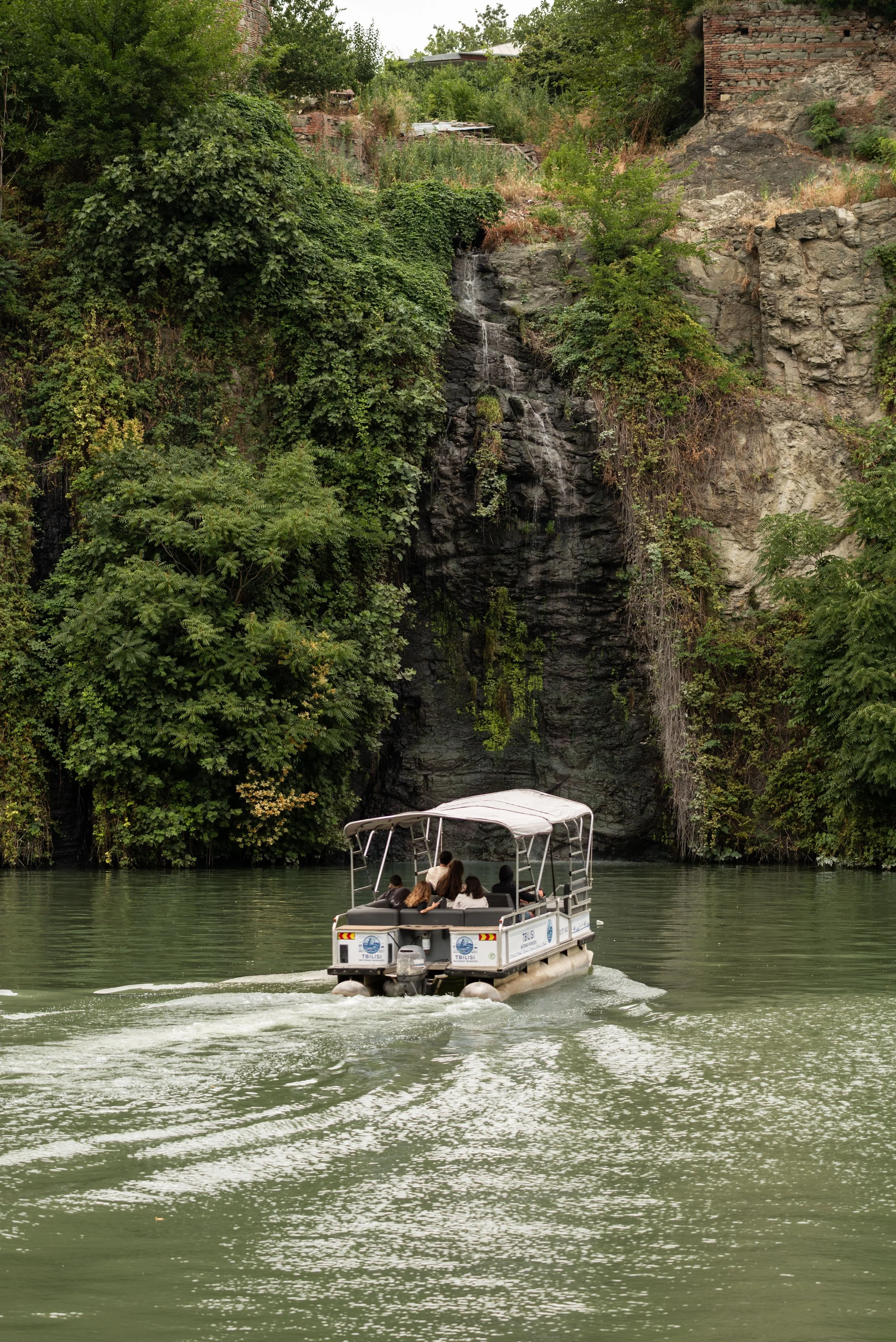 A boat with four people on a river near green trees and a rocky cliff with some buildings at the top, and a small waterfall.