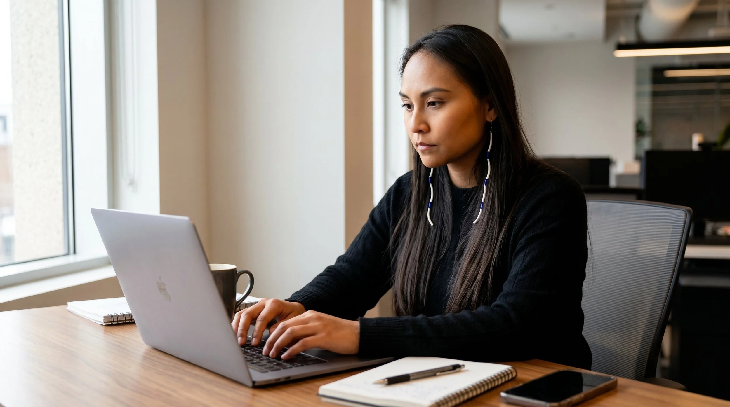 A woman with long dark hair wearing a black sweater and earphones working on a laptop at a desk in a modern office space with large windows.