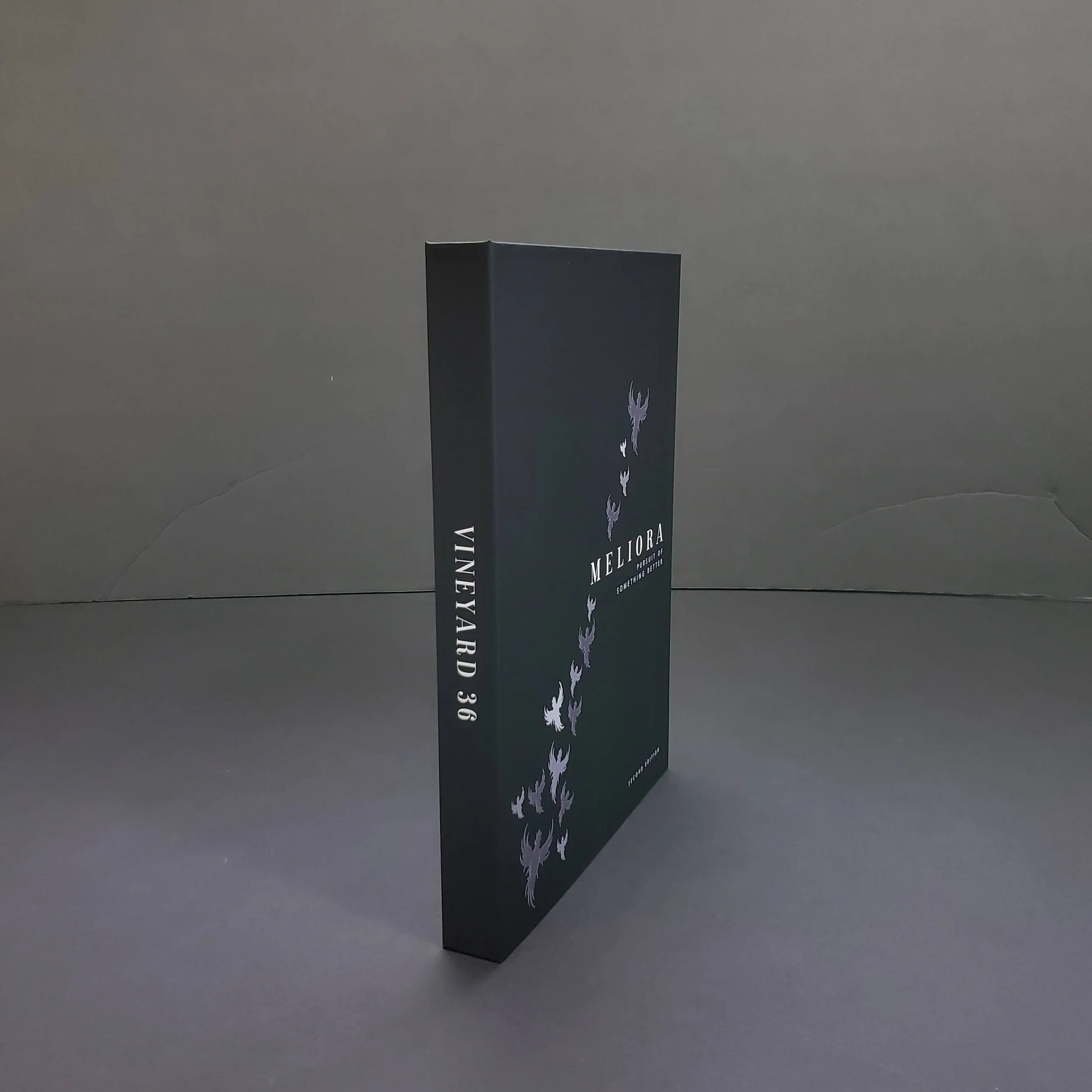 A tall black rectangular box with white floral patterns and the text 'MELIORA' written vertically on one side, placed on a flat grey surface against a plain grey background.