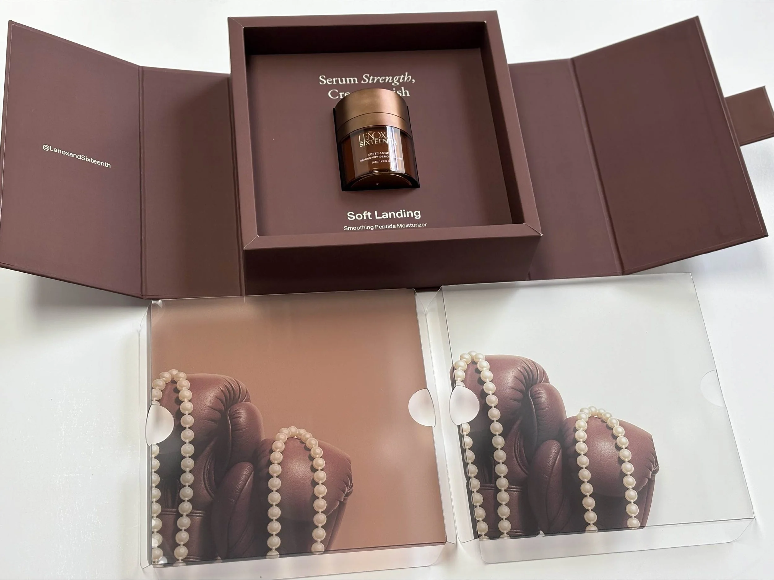 A brown box containing a small jar of serum labeled "LENOX SIXTEEN" with a gold lid, and the words "Soft Landing" and "Smoothing Peptide Moisturizer" on the label, with a background featuring a magazine or advertisement displaying jewelry and a leath