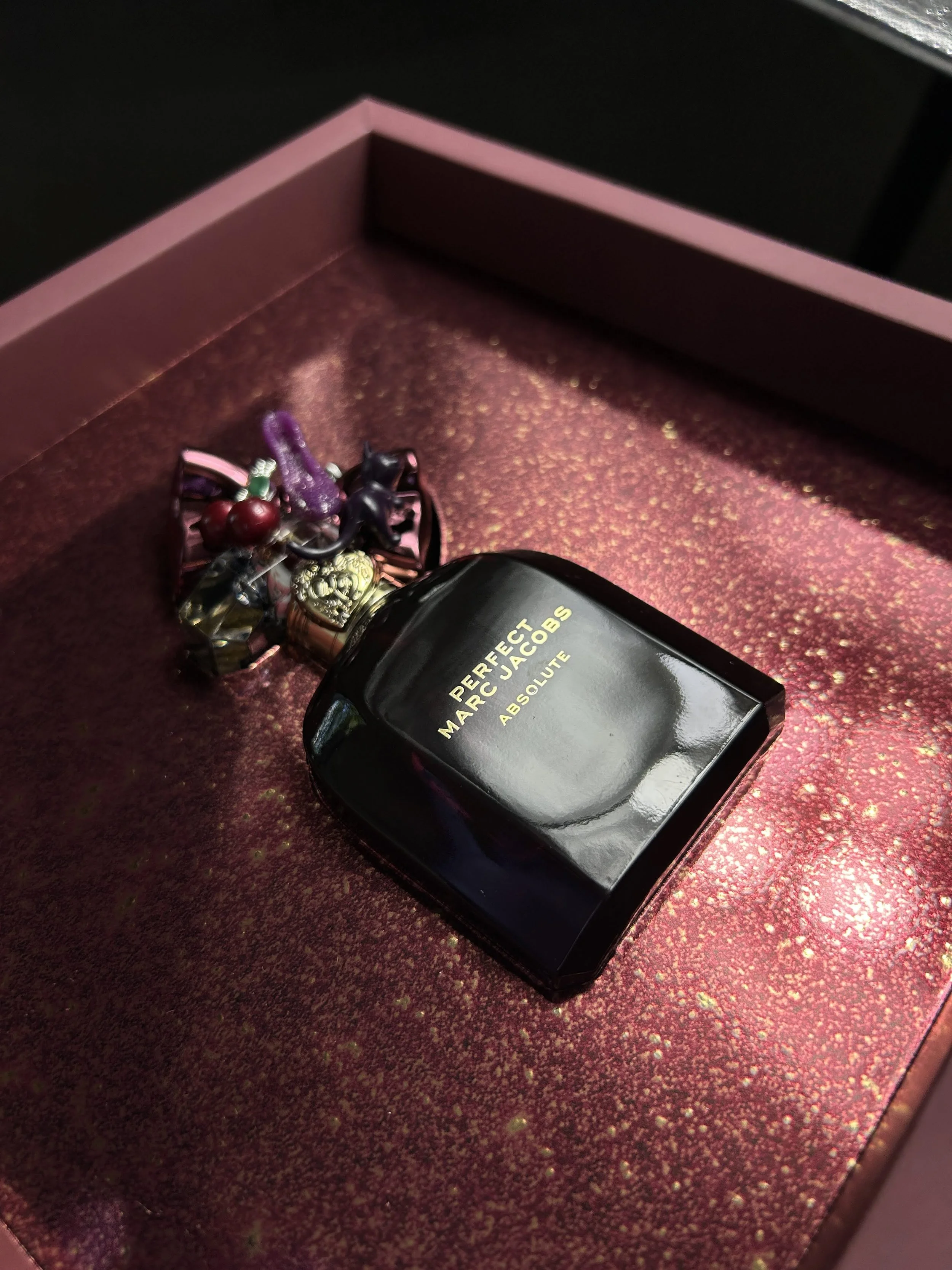 A black bottle of Marc Jacobs perfume labeled 'PERFECT' and 'ABSOLUTE' is placed on a glittery, red-speckled surface inside a pink and black box filled with assorted jewelry, including rings, bracelets, and a small green plastic object.