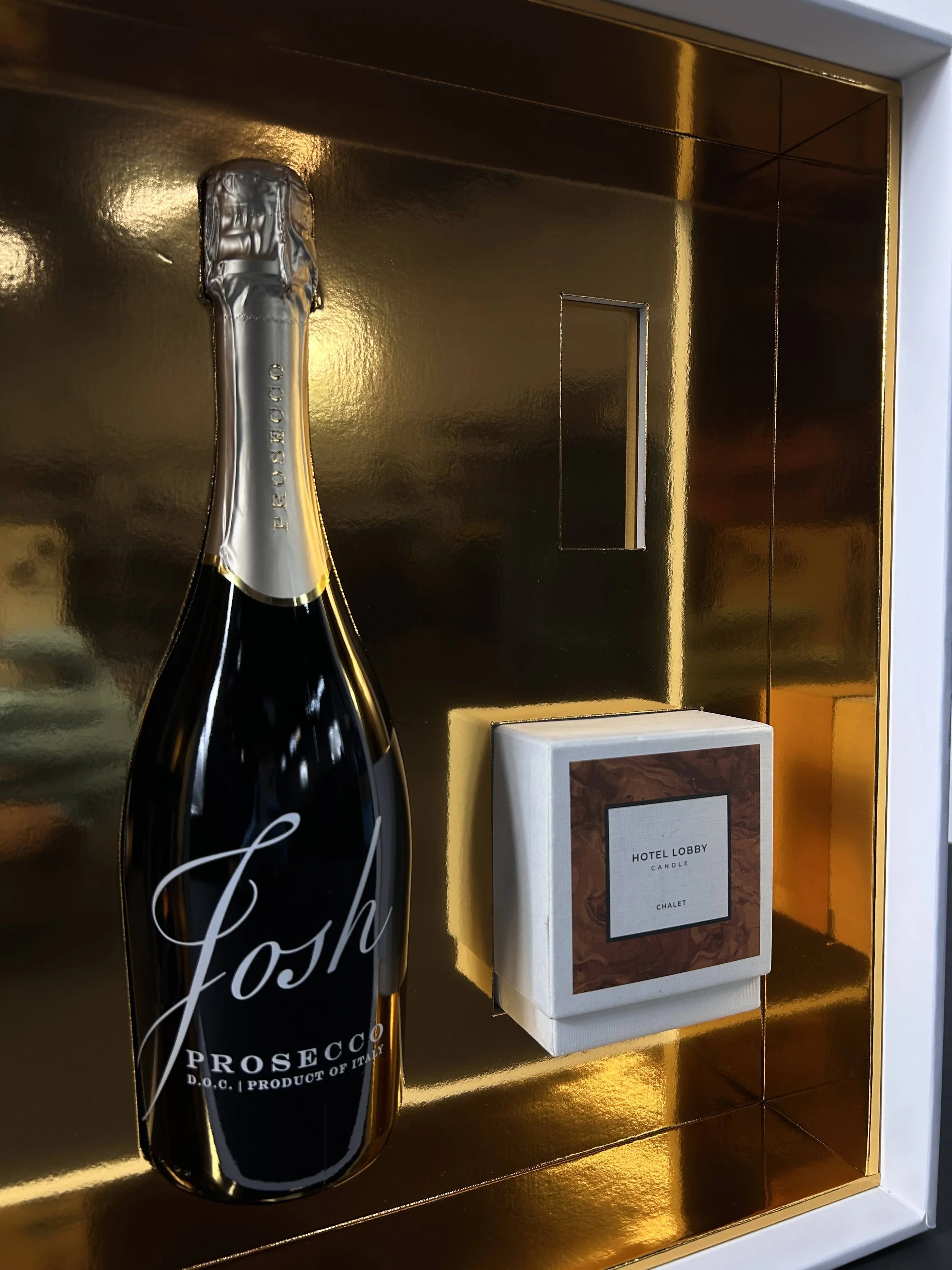 A bottle of Prosecco champagne with a gold foil top, positioned against a golden wall, alongside a boxed candle labeled "Hotel Lobby Candle Chalet".