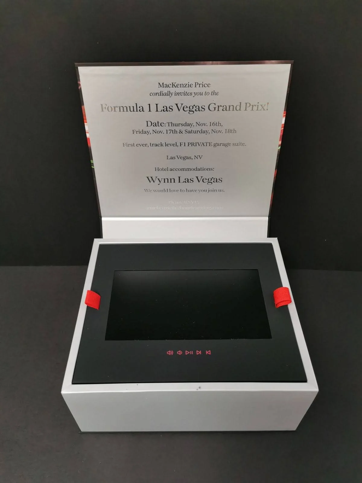 A black electronic device with a blank screen and red control icons below the screen, placed in a white box with red straps, in front of a black background, with a printed invitation for the Formula 1 Las Vegas Grand Prix event behind it.