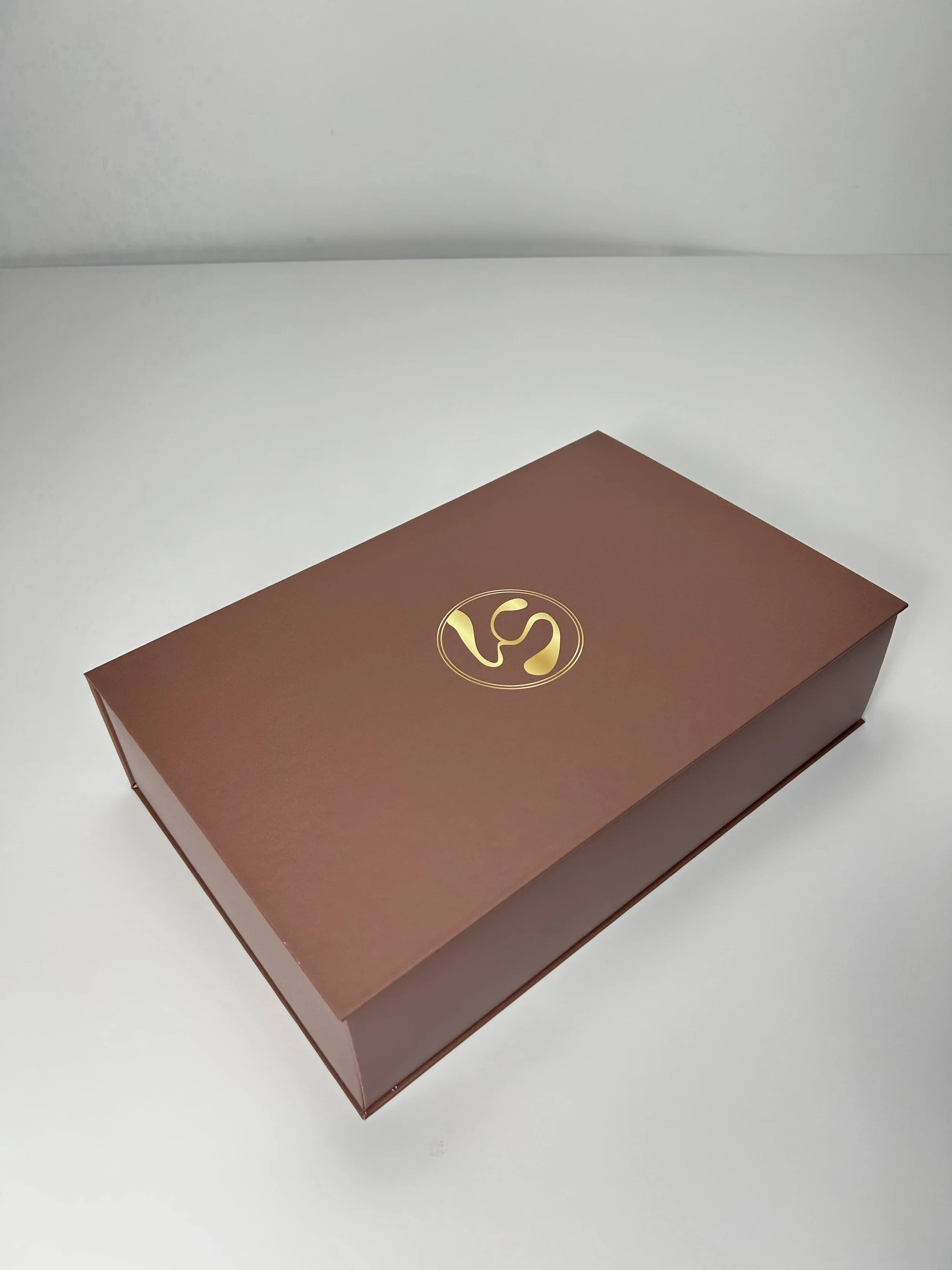 A rectangular brown box with a gold circular logo on the top, placed on a white surface against a white background.