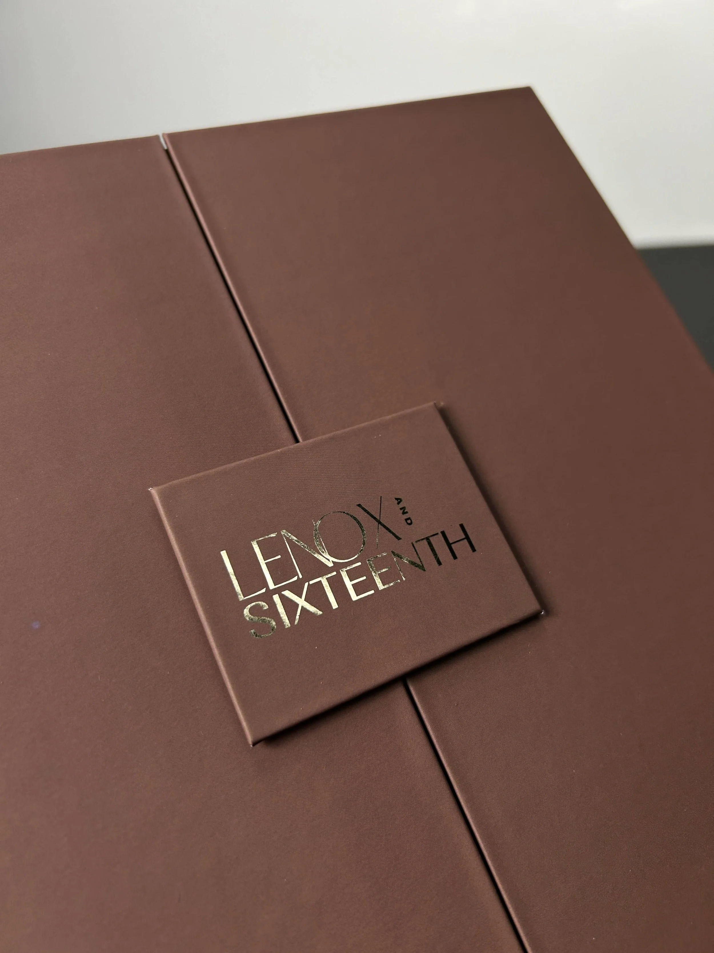 Close-up of a pinkish-brown box with silver foil text that reads 'LENOX AND SIXTEENTH' on a matching pinkish-brown surface.