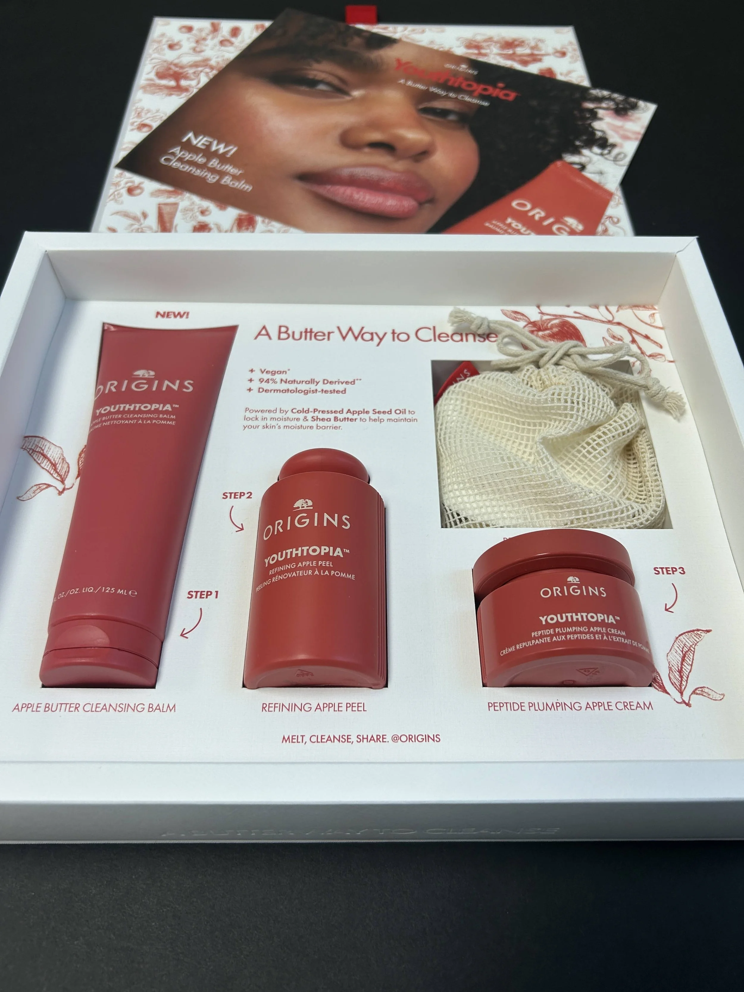 Packaging box for Origins skincare products featuring a large image of a woman with natural curly hair. Inside the box are three skincare products: a cleansing balm, an apple peel, and an apple cream, all in red containers. The box and products empha