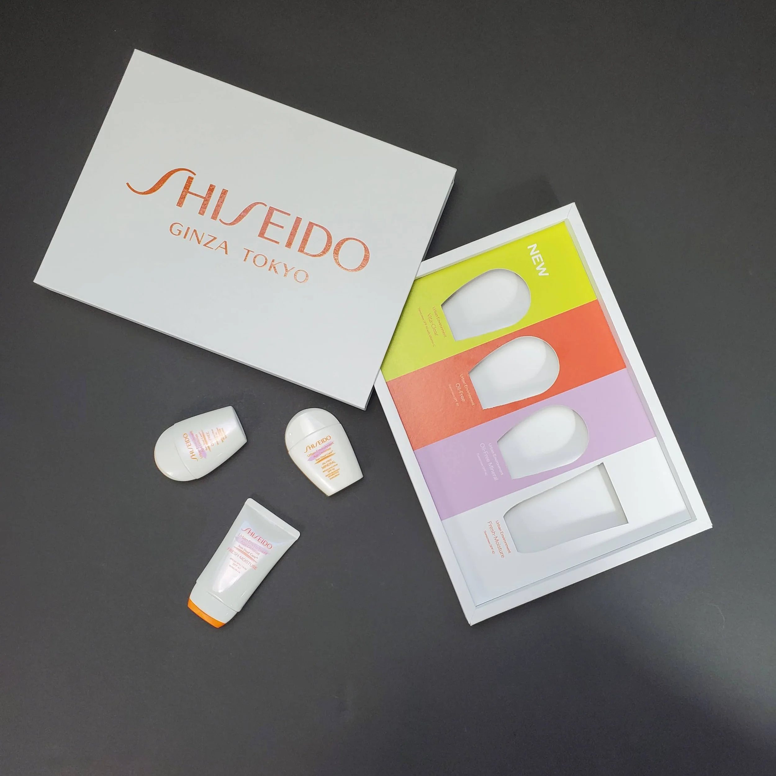 Assorted skincare product packaging from Shiseido on a black surface, including a white box, test tube bottles, and colorful sample sachets.