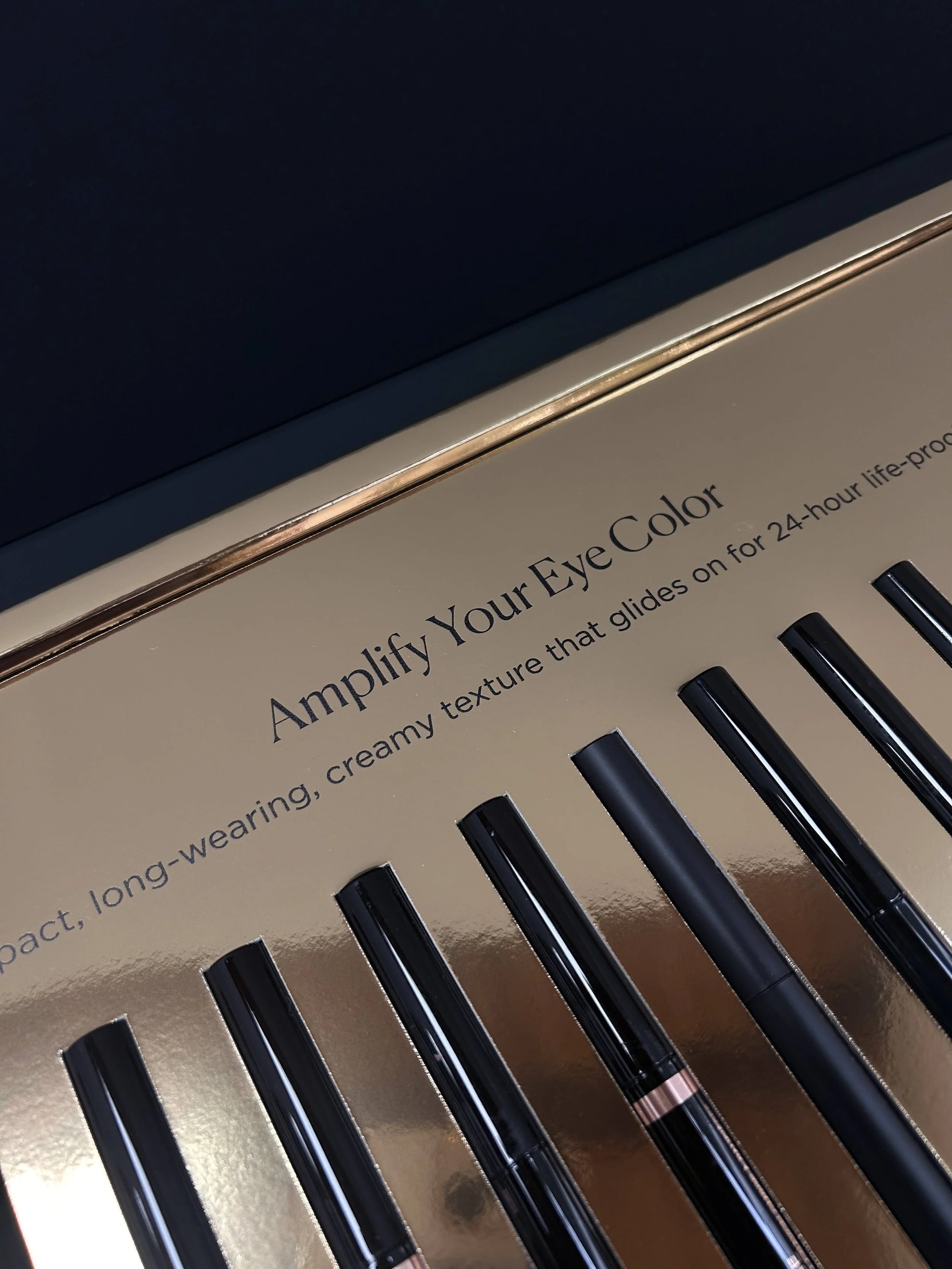 Close-up of a makeup palette with black eyeliner pencils in front and the palette text partially visible, reading 'Amplify Your Eye Color.'