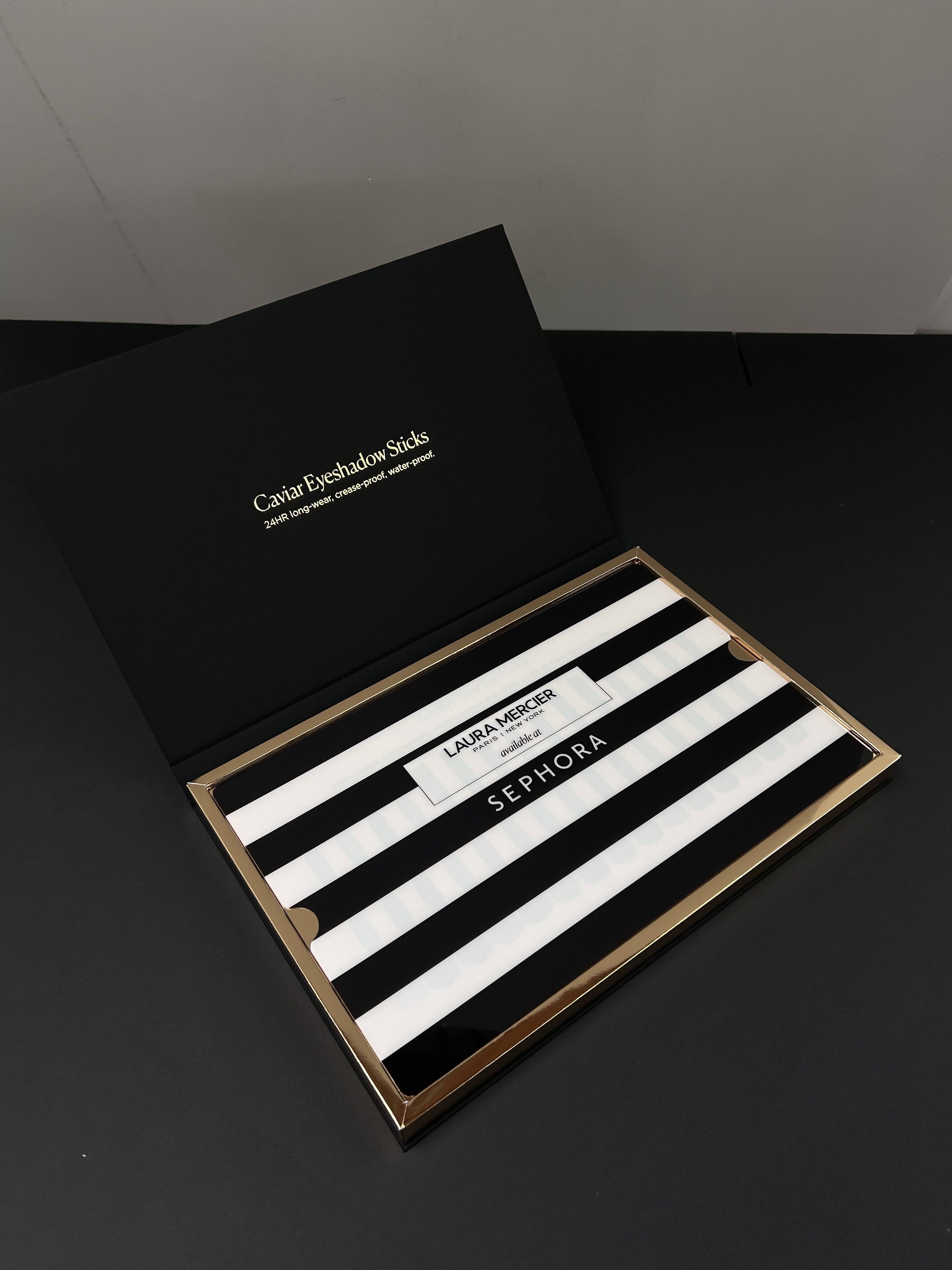 Open black box labeled 'Caviar Eyeshadow Sticks, 24HR long-wear, crease-proof, water-proof' with black and white striped eye shadow sticks inside, placed on a black surface.