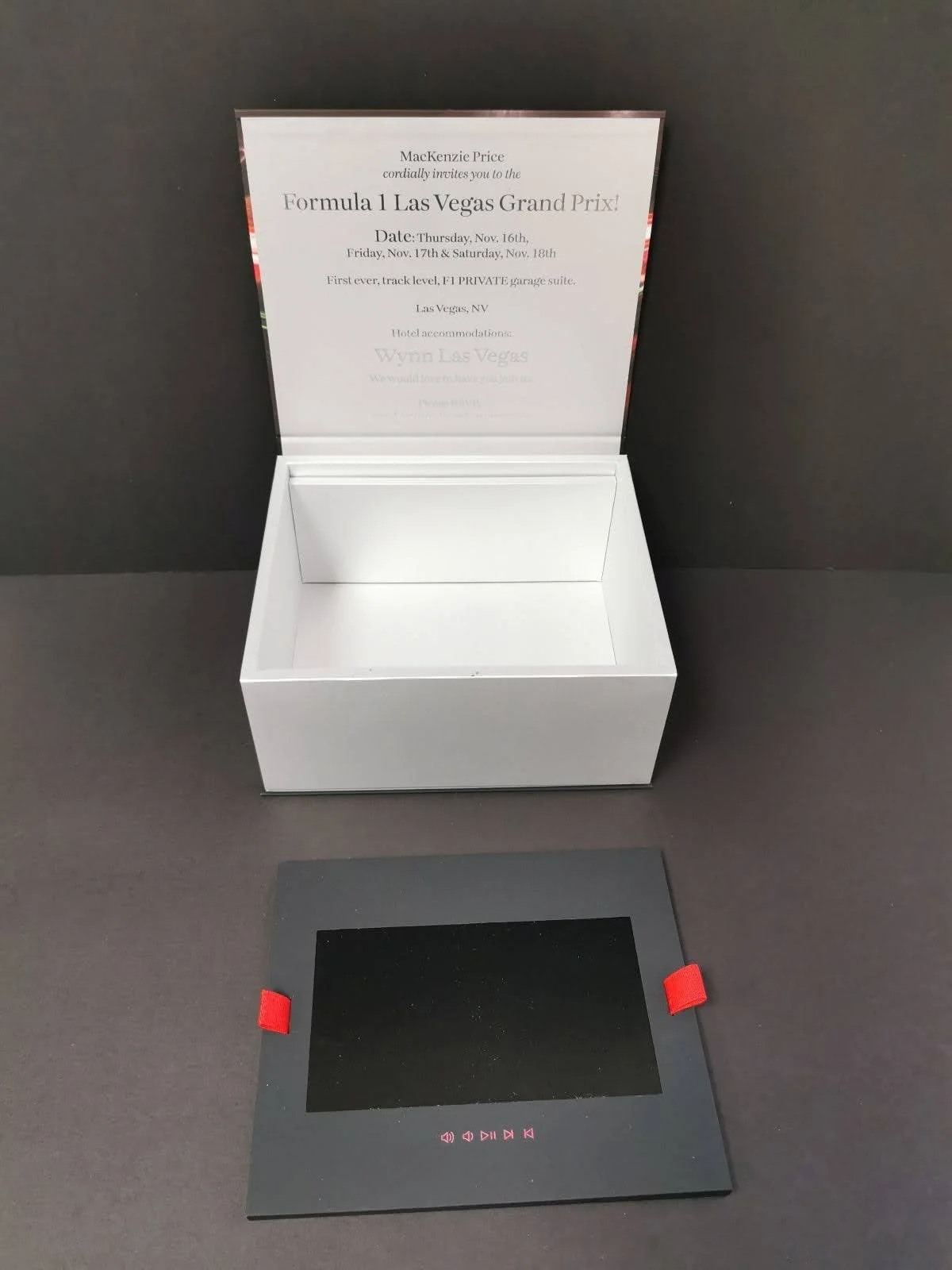 A white box with a printed invitation inside, and a black electronic device with red accents on a dark surface.