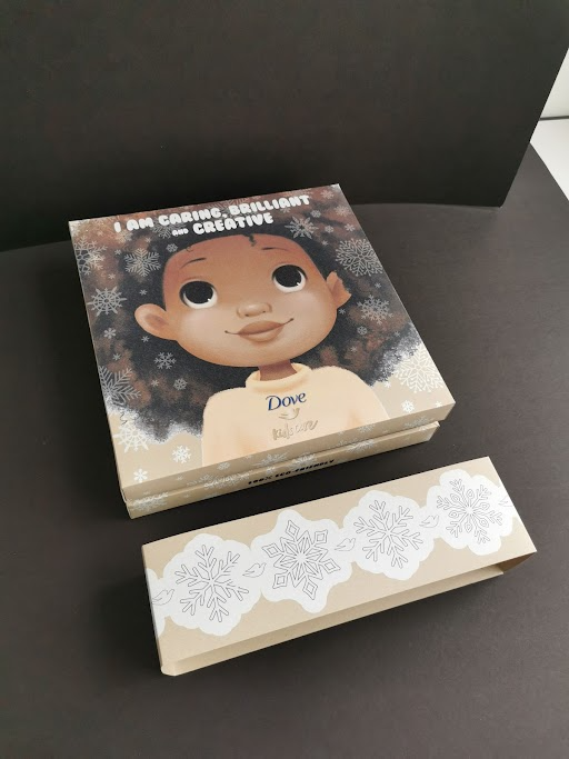 A stack of cards featuring a cartoon girl with dark skin, big eyes, and curly hair, surrounded by snowflakes, with the words "I am caring, brilliant, and creative" at the top, placed on a black surface.