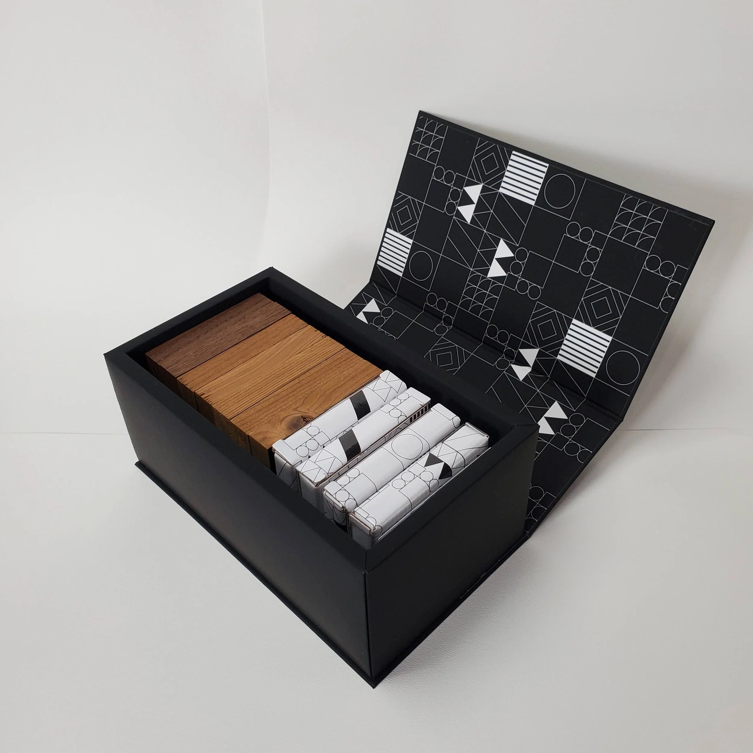 A black box with a geometric pattern containing white shapes, partially opened to reveal several wooden planks and patterned wrapping papers inside, situated on a white surface against a white wall.