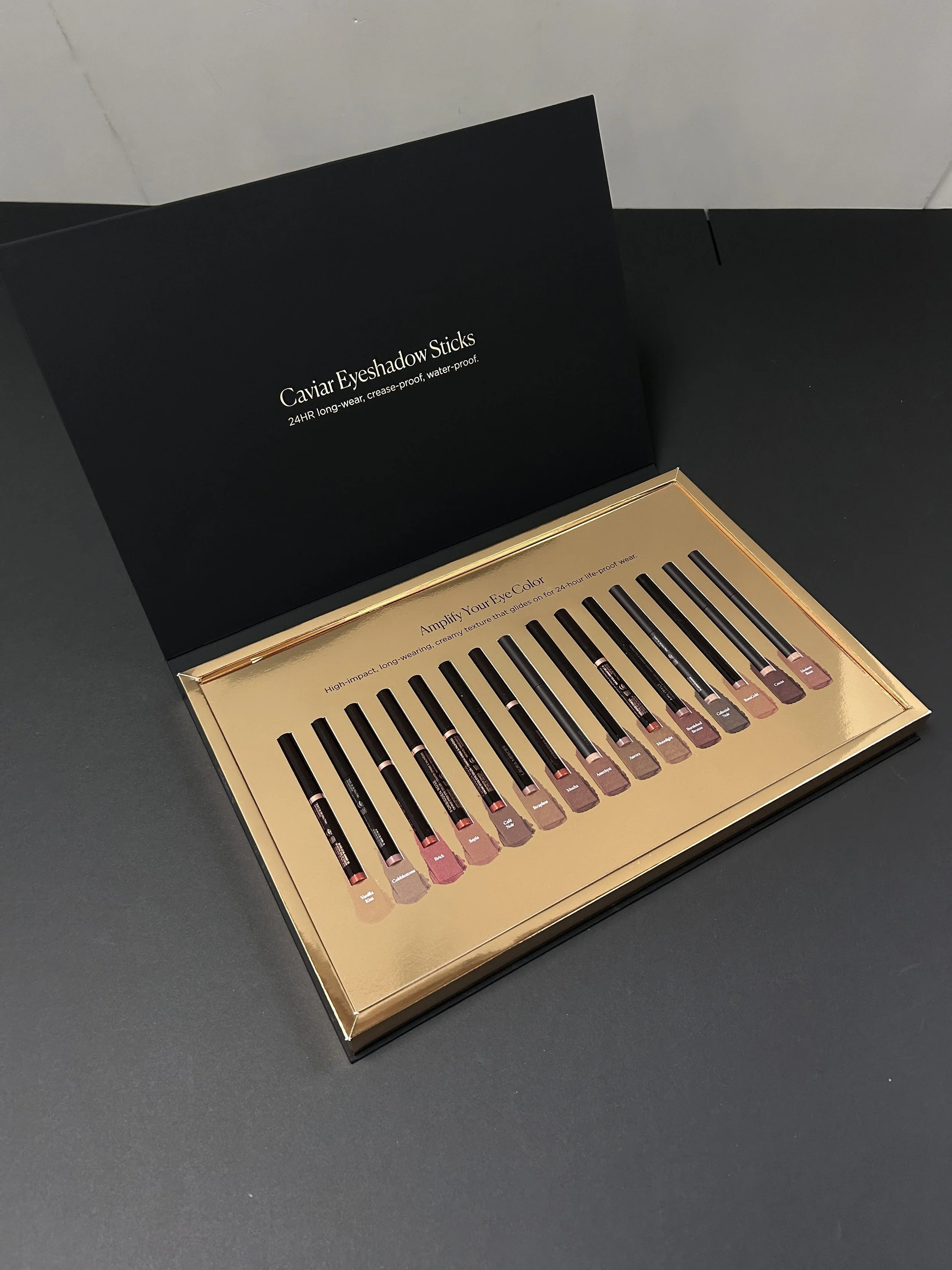 Open box of Caviar Eyeshadow Sticks with multiple shades displayed inside, with black packaging and gold accents.