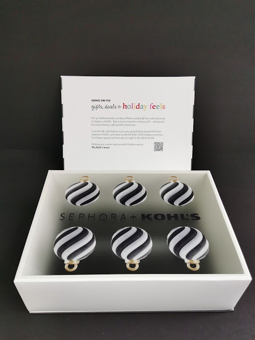 Six black and white striped Christmas ornament decorations inside a white box with 'SEPHORA+KOHL'S' printed inside, and a card with holiday message behind the decorations.