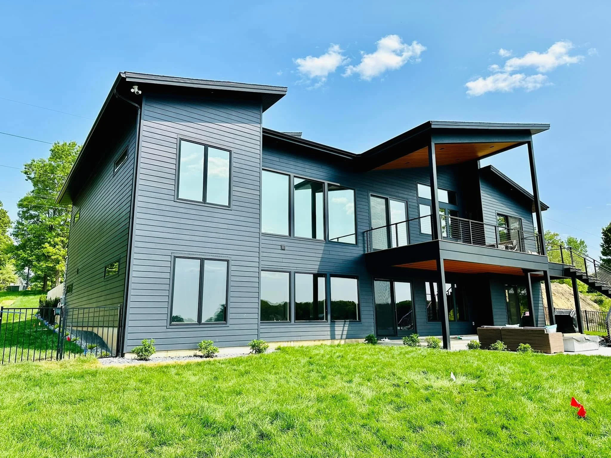 Modern multi-story house with blue exterior, large windows, and a balcony overlooking a grassy yard under a blue sky with scattered clouds.