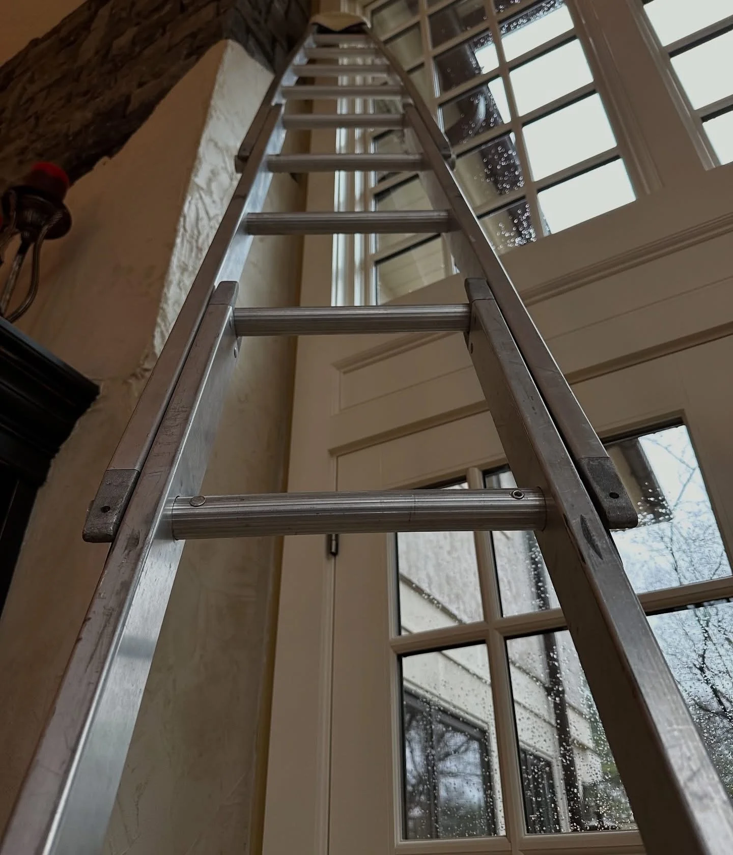 An aluminum ladder extending inside a room with large window panes and a brick and white wall. The room has a view of a snowy outdoor scene with trees.