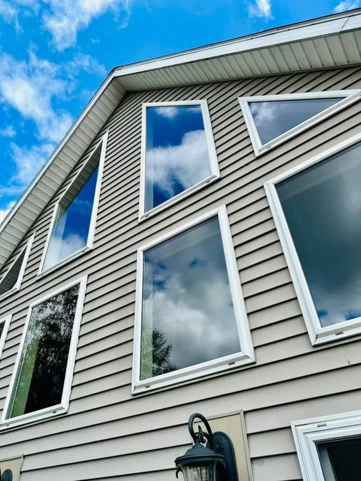 View of a modern house with beige siding and multiple windows reflecting a blue sky with clouds, seen from below.