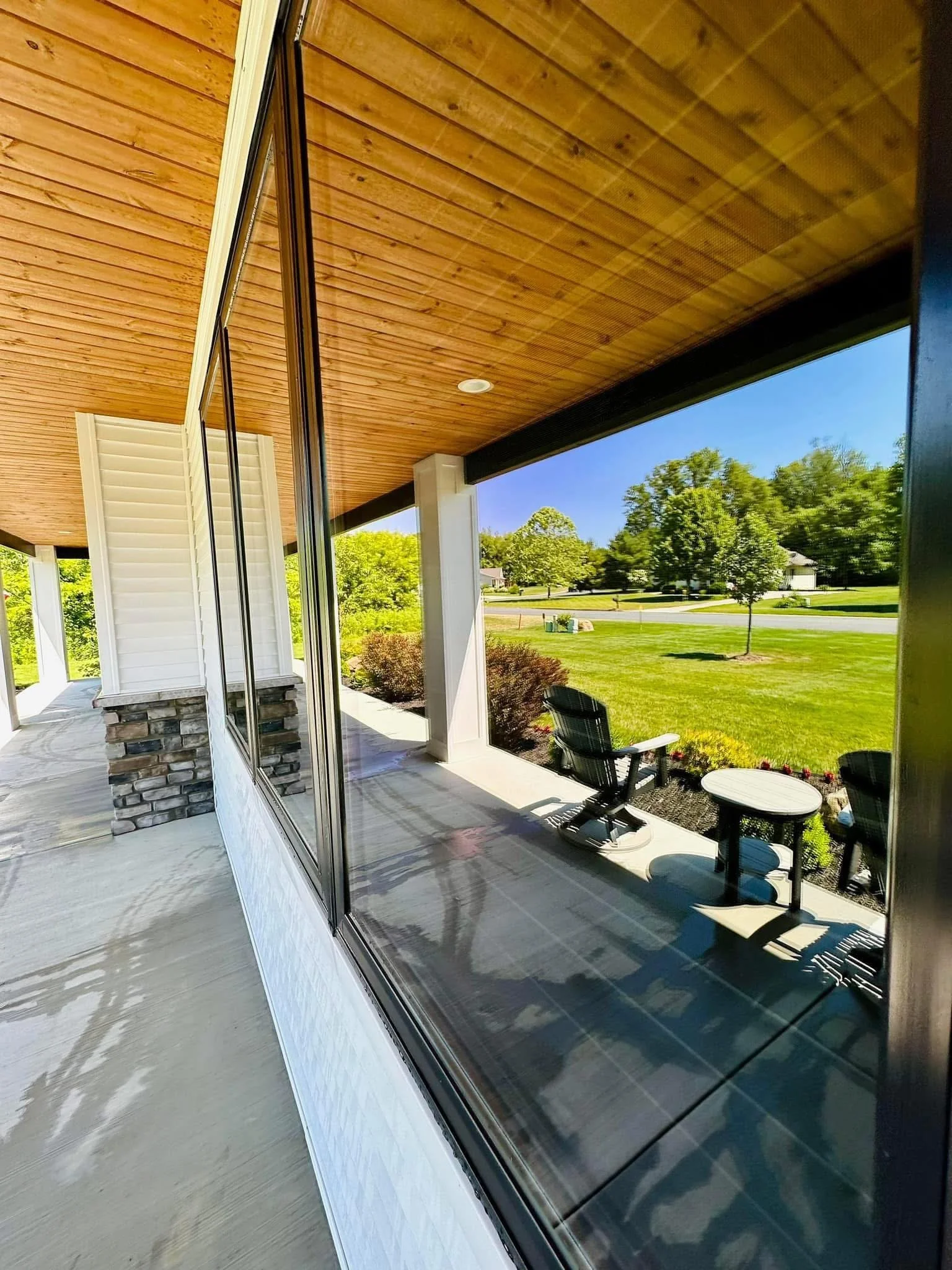 View from porch showing a glass sliding door, outdoor patio furniture, green lawn, trees, and houses in the distance under a clear blue sky.