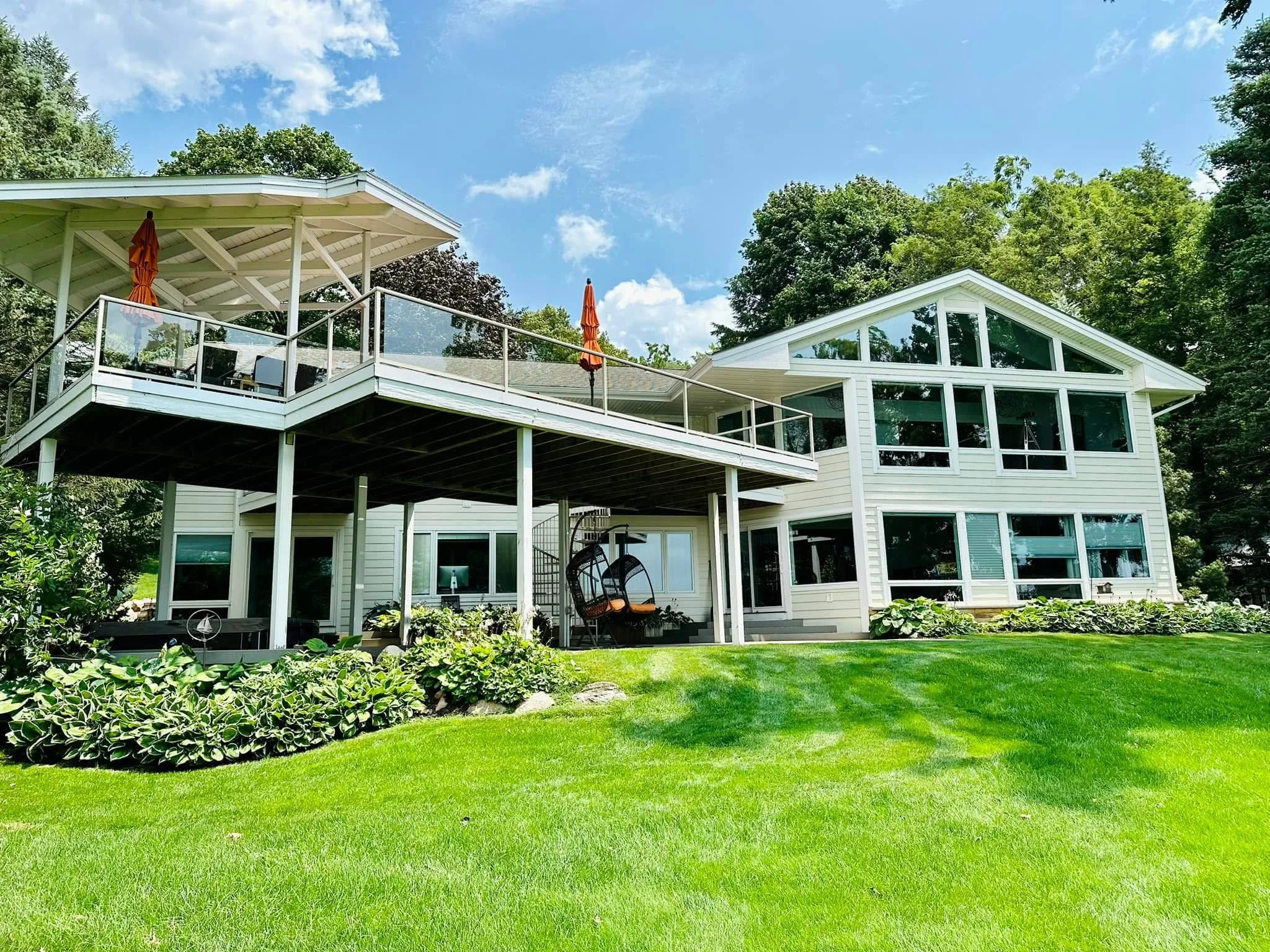 A modern white house with large windows and a spacious deck, surrounded by lush greenery and a well-maintained lawn under a blue sky with scattered clouds.