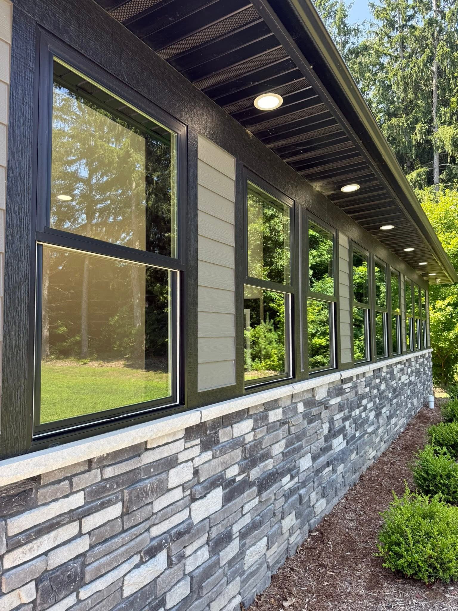 Exterior view of a house with large windows, black trim, and stone foundation, surrounded by greenery.