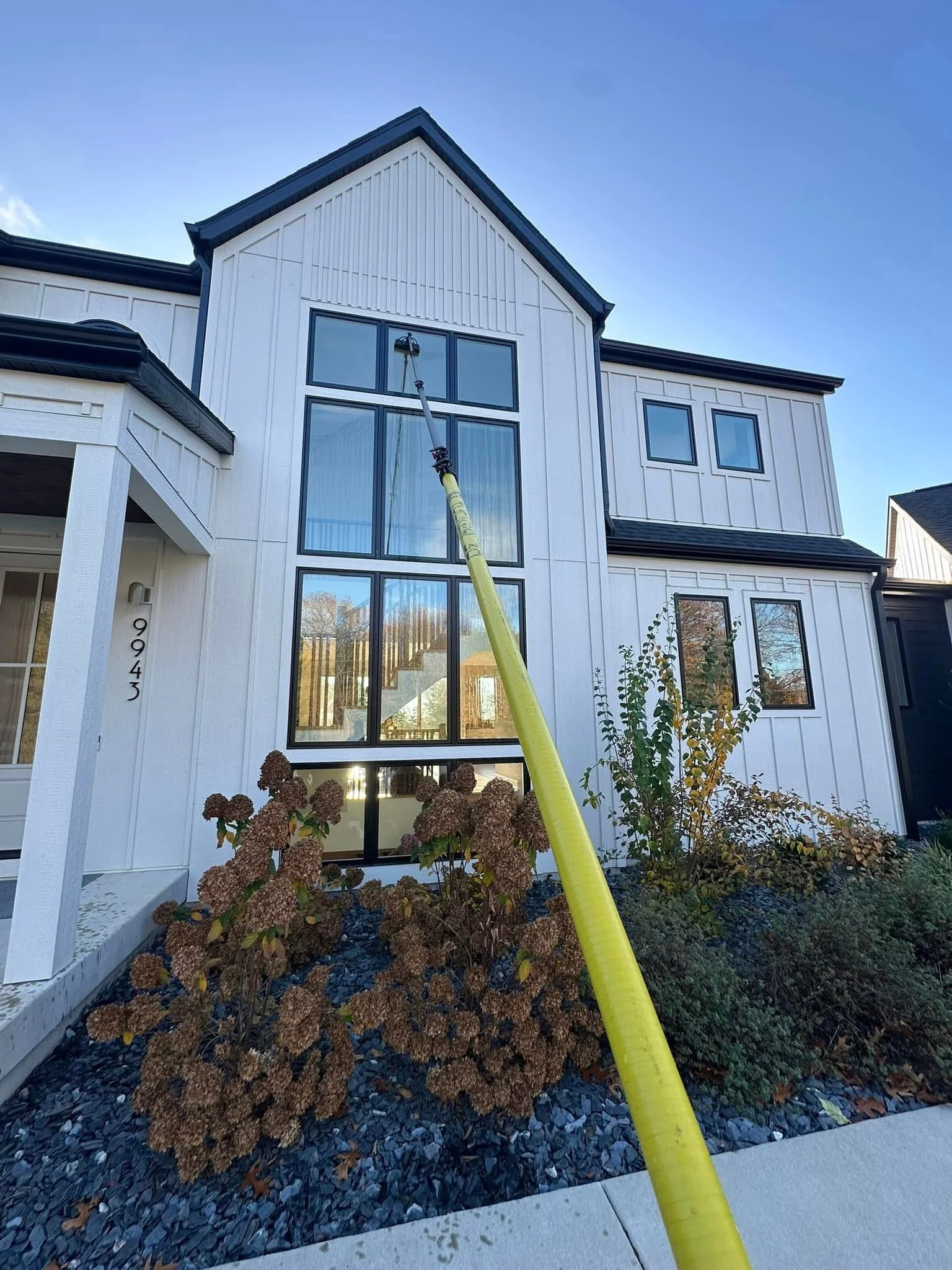 Front of a white modern house with large windows and a garden bed with dried brown plants, being cleaned with a long pole attachment during daytime.