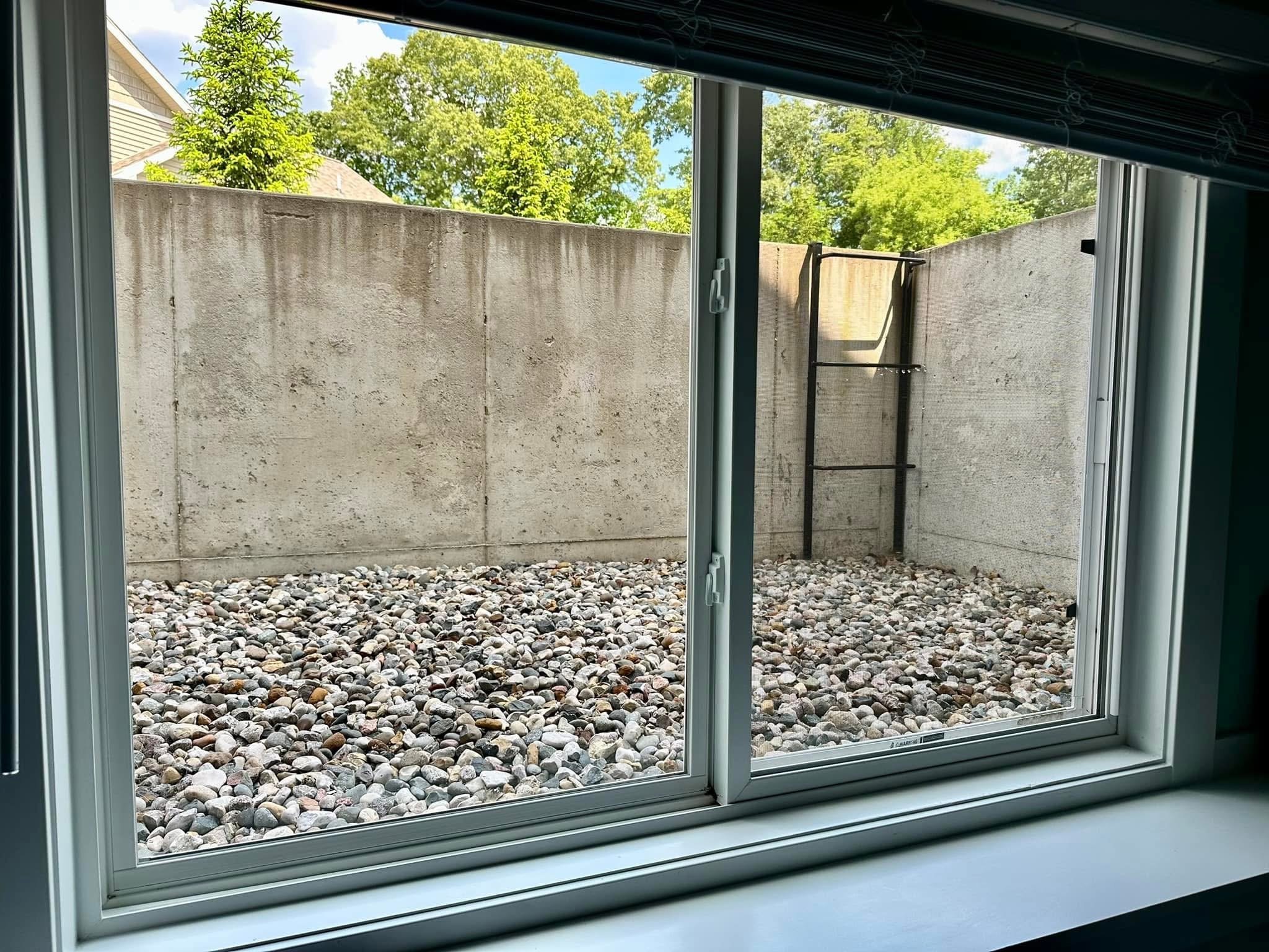 View through a sliding glass door showing a backyard with a gravel surface, a tall concrete fence, green trees, and a partly cloudy sky.
