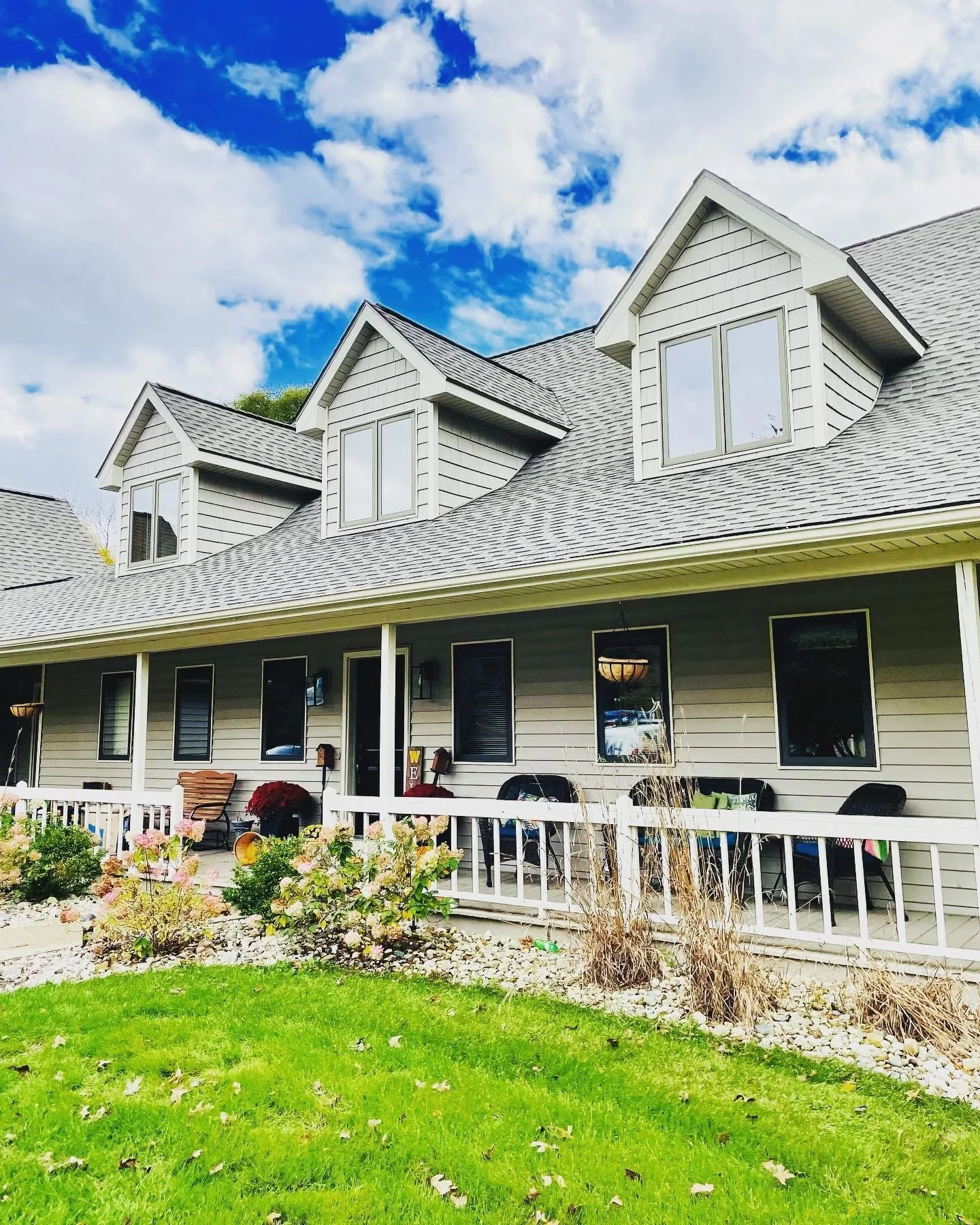 A two-story house with gray siding and a gabled roof, featuring three dormer windows. The house has a porch with outdoor furniture, potted plants, and a white picket fence in a yard with green grass, flowers, and dried plants, under a partly cloudy s