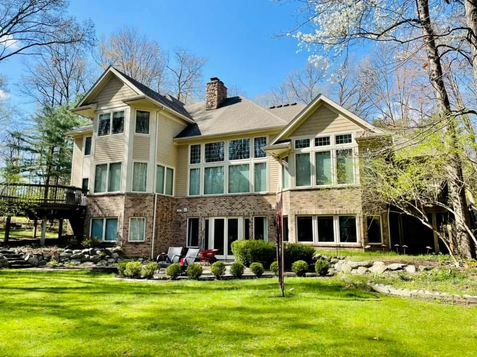 Large two-story house with beige siding, brick foundation, and numerous large windows, surrounded by a well-maintained lawn, trees, and a clear blue sky.