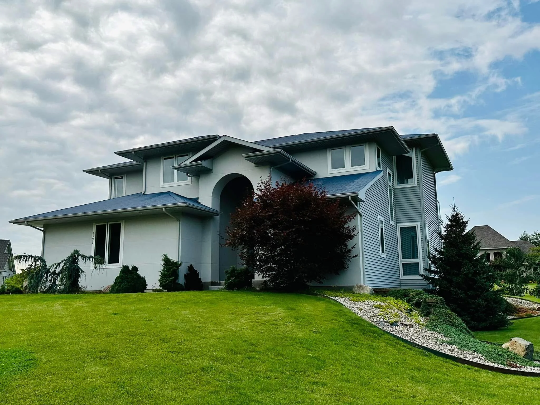 A modern, multi-story house with a well-kept green lawn and landscaped garden, including small bushes and a tree, under a partly cloudy sky.