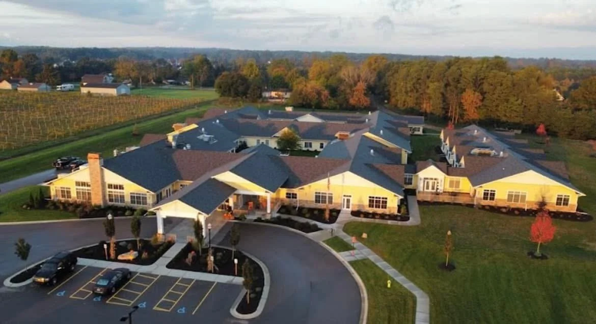 A large residential complex with multiple connected yellow buildings, a parking lot with several cars, surrounded by green lawns, trees in fall colors, and a vineyard in the background.
