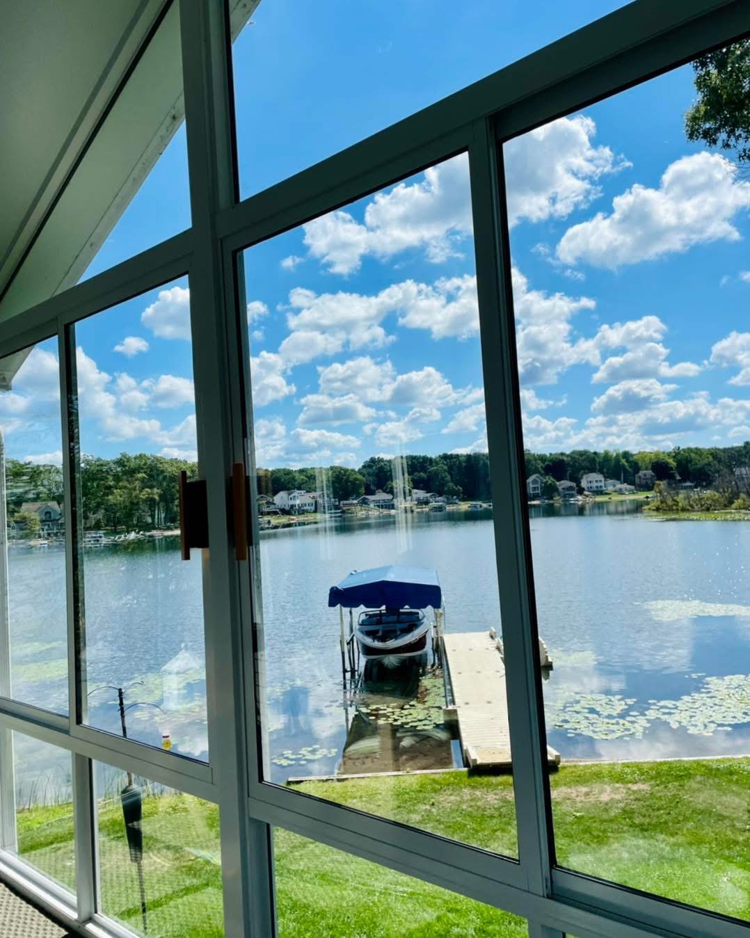 View of a boat docked at a lakeside from inside a building with large glass windows, reflecting the blue sky and white clouds.