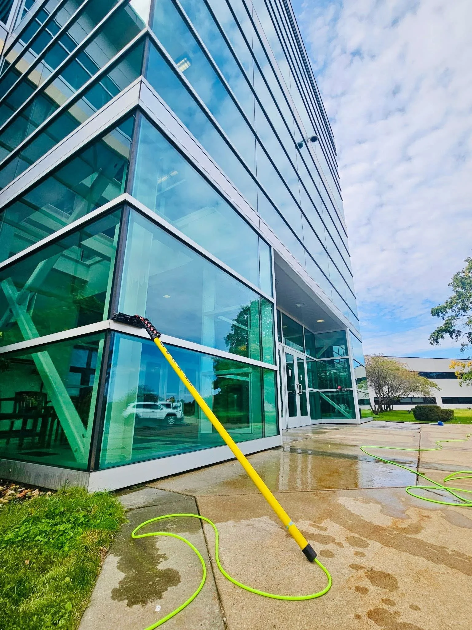 Exterior view of a modern glass office building with wet concrete and a green hose on the ground, indicating recent cleaning or maintenance.