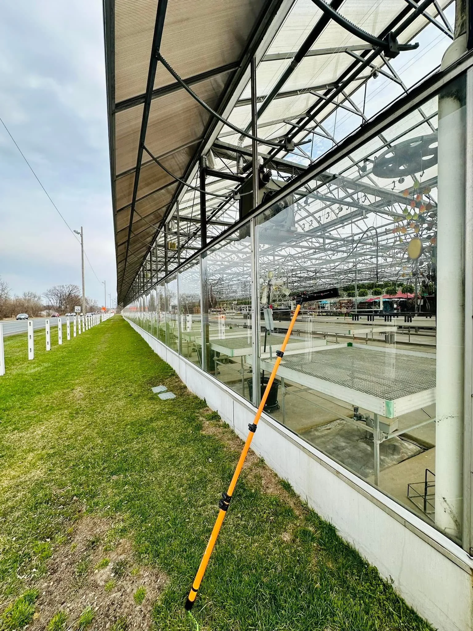 View of a greenhouse with glass walls and metal framing, located beside a road. Outside, the grass is green, and there are power lines and cars on the road. Inside the greenhouse, gardening equipment and structures are visible. An orange and black po