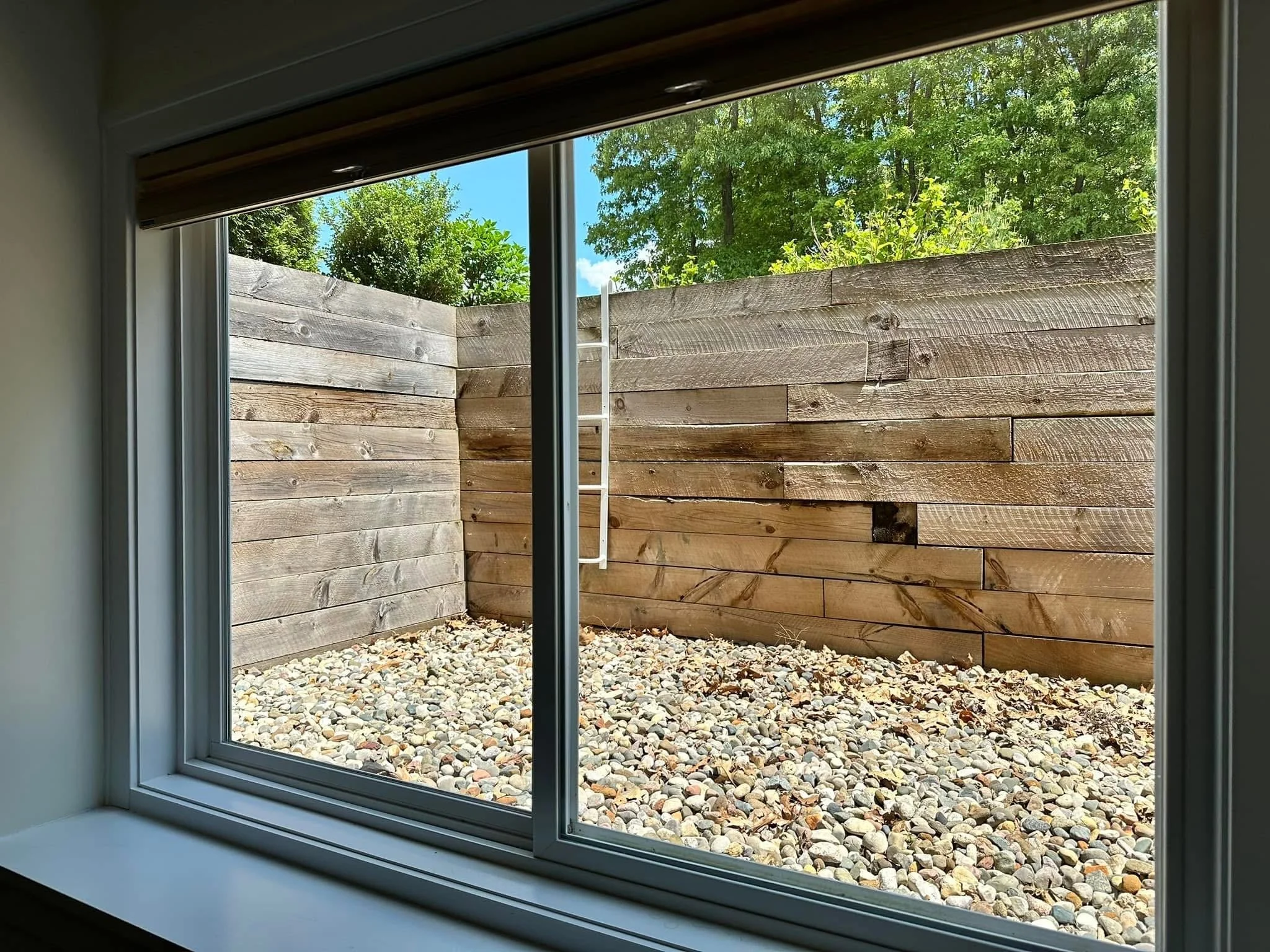 View of a wooden fence outside a window with a gravel ground and green trees in the background.