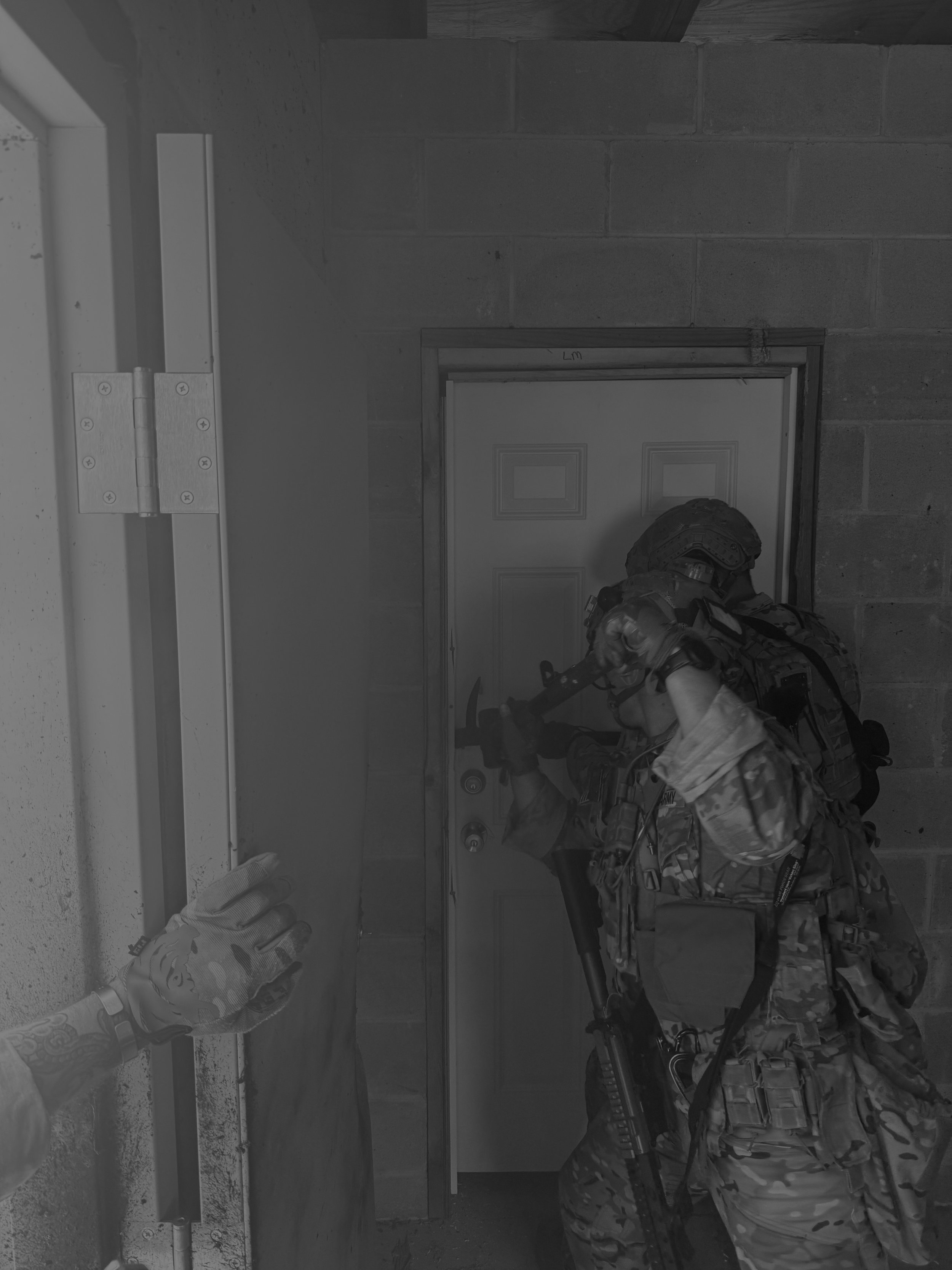 A soldier in camouflage uniform and helmet sitting against a wall in a room with concrete walls, holding a rifle, with a person’s arm extended holding a white ring. special operators conducting a breach on a door