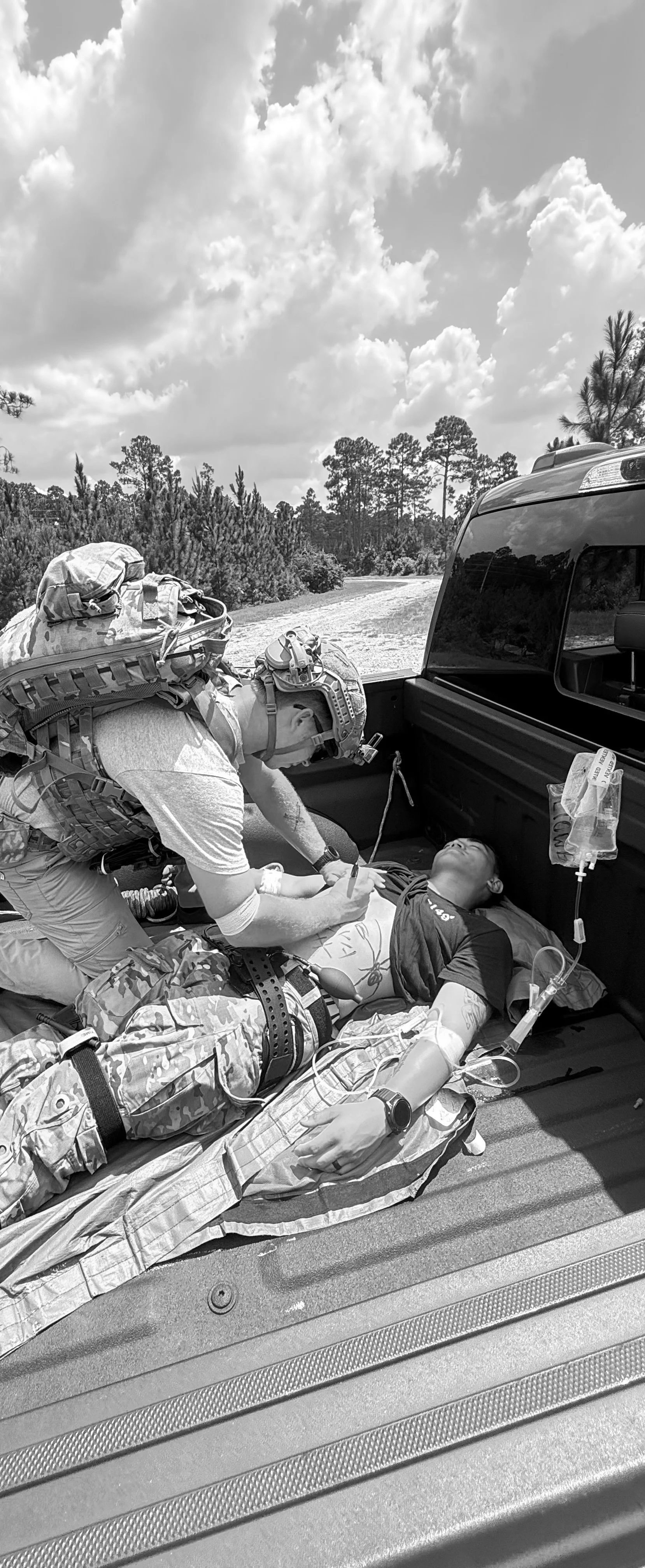 A soldier receiving medical aid from a responder on a military-style vehicle bed outdoors, with trees and cloudy sky in the background.
special operations medic giving aid