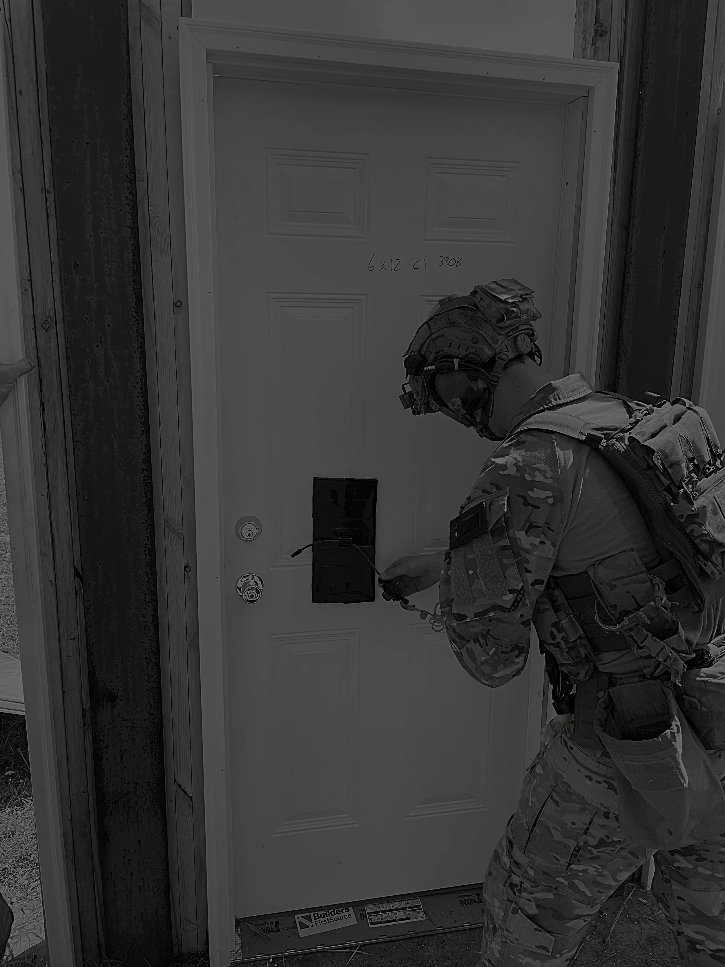 A soldier in camouflage uniform with tactical gear and a helmet inspecting a white door with a frame while placing explosives on the door for a breaching charge.