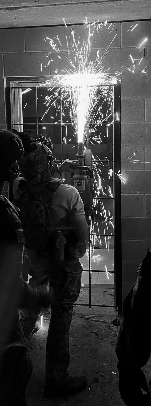 Soldiers in tactical gear practicing breaching a reinforced door with a mechanical saw, sparks flying from the cutting process.