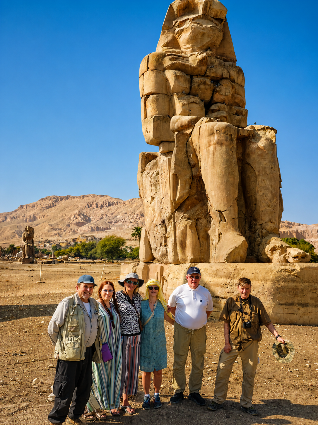 Group of tourists standing in front of a tall ancient stone statue of King Ramses II at a historical site in Egypt, with desert landscape and mountains in the background.
