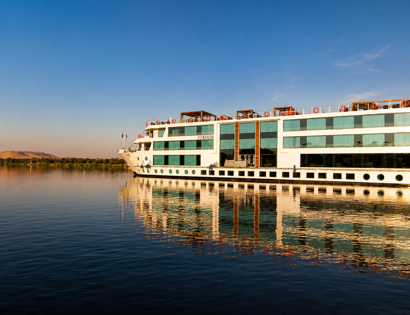 A large white cruise ship named Esmeralda floating on calm river water during sunset, with trees and hills in the background.