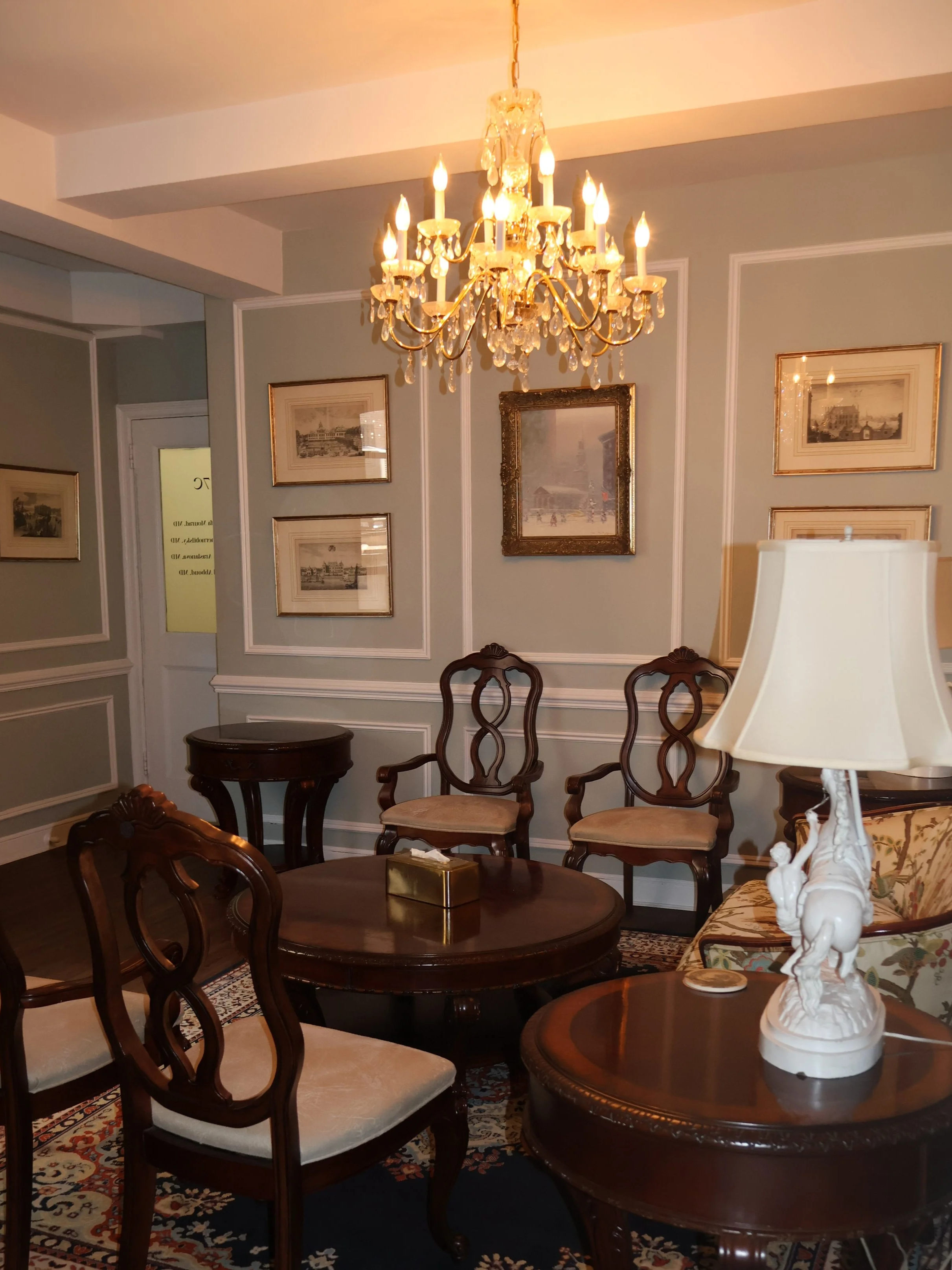 An elegant living room with a crystal chandelier, framed artwork on light gray walls, a dark wooden dining table with matching chairs, a decorative lamp on a side table, and an ornate white sculpture on a side table.